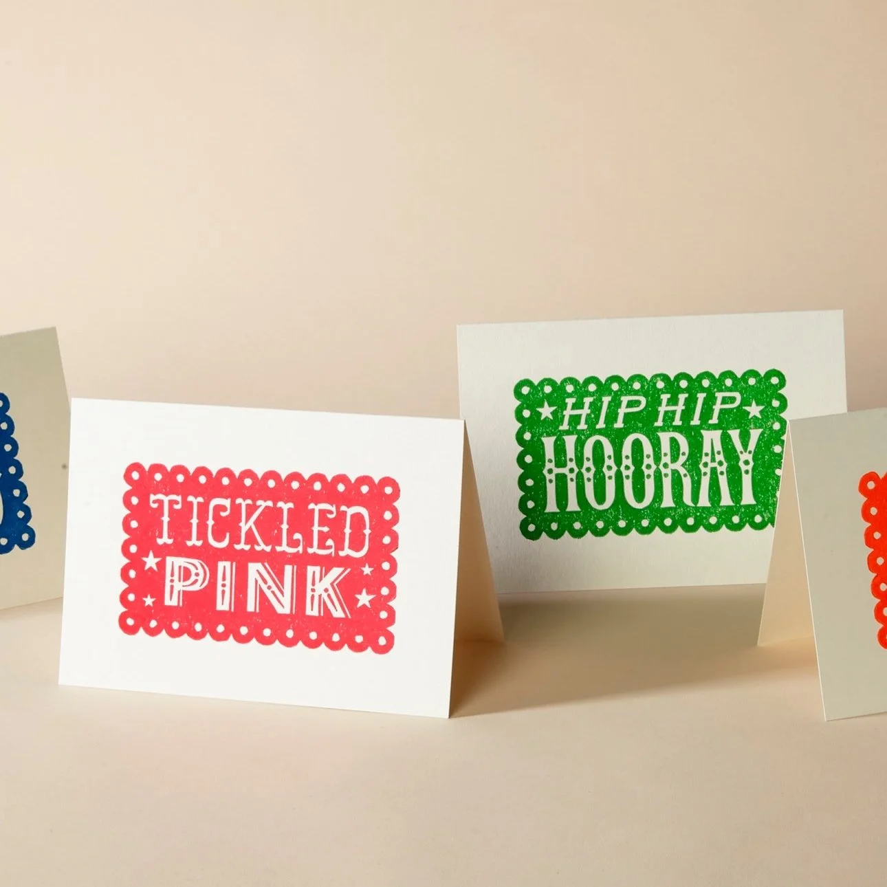 Four greeting cards with colorful designs and cheerful phrases like "Tickled Pink" in red and "Hip Hip Hooray" in green, against a neutral background.