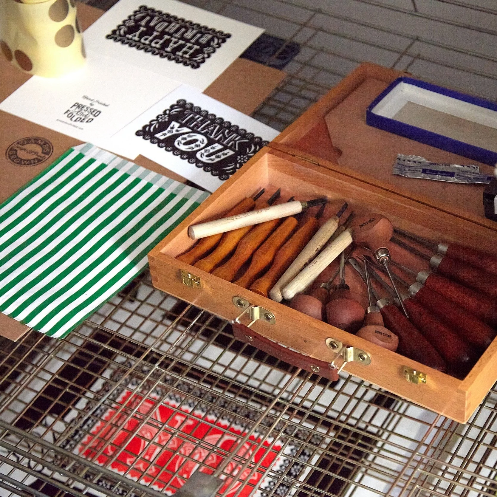 Wooden case with carving tools on a wire rack, alongside printed cards and green-striped bag.