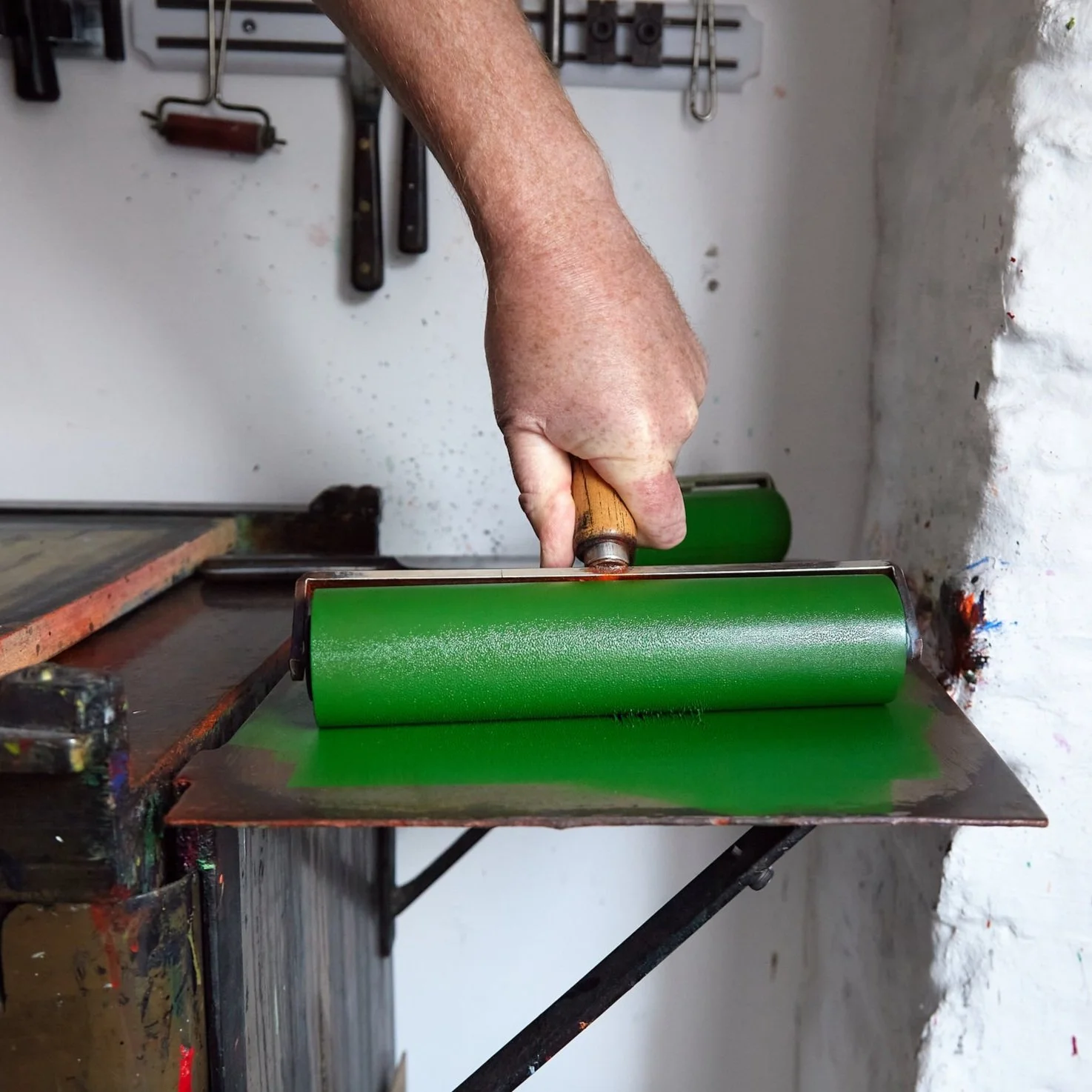 A person using a brayer or roller to apply green ink on a surface, likely for printmaking, with tools hanging on the wall in the background.