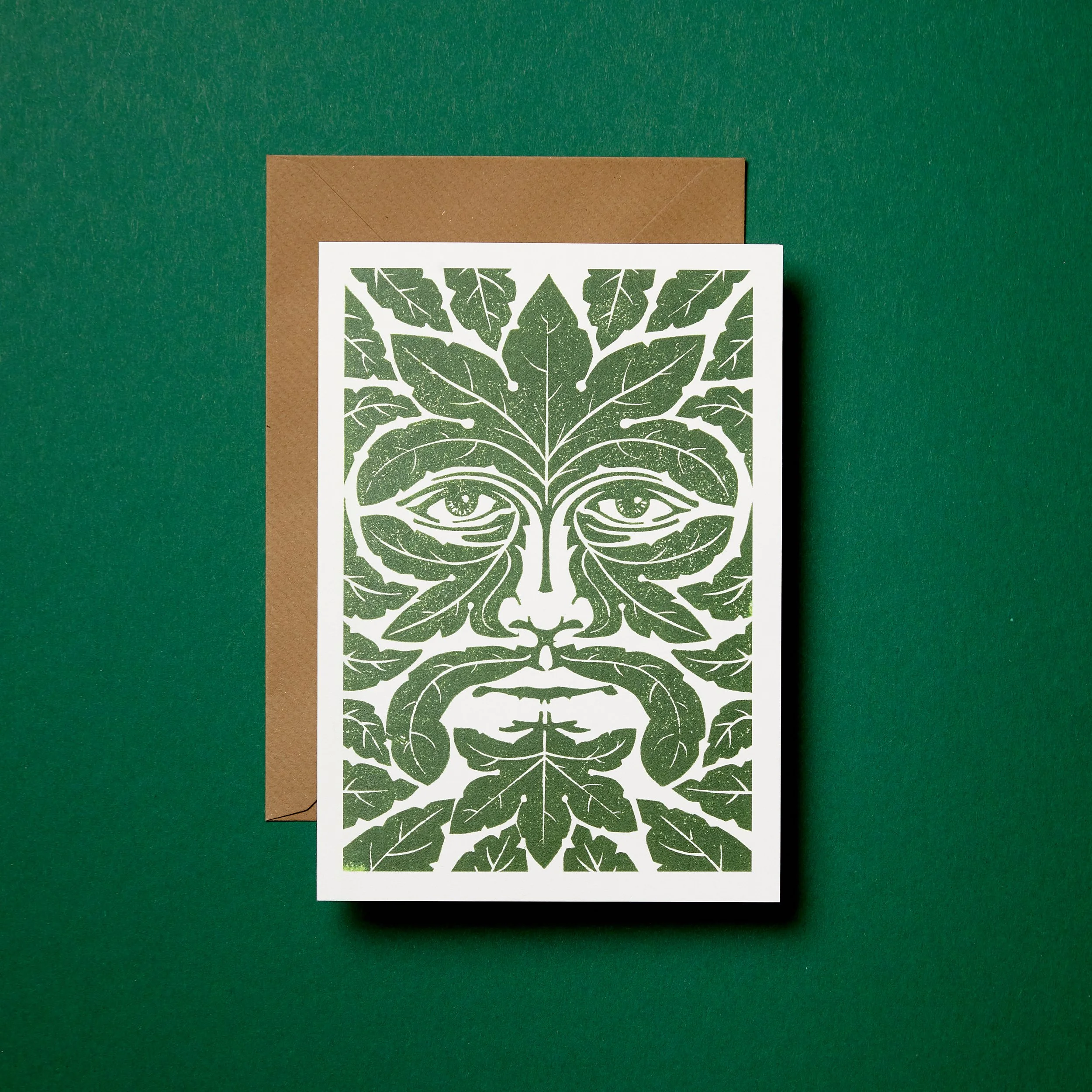 Greeting card featuring a Green Man design with a symmetrical face made of leaves on a green background, accompanied by a brown envelope.