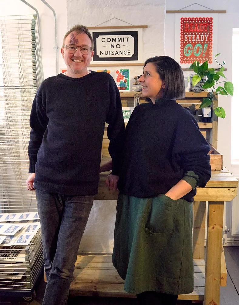 Two people standing smiling in a studio by a table showing linocut COMMIT NUISANCE and READY STEADY GO! With potted plant and drying rack