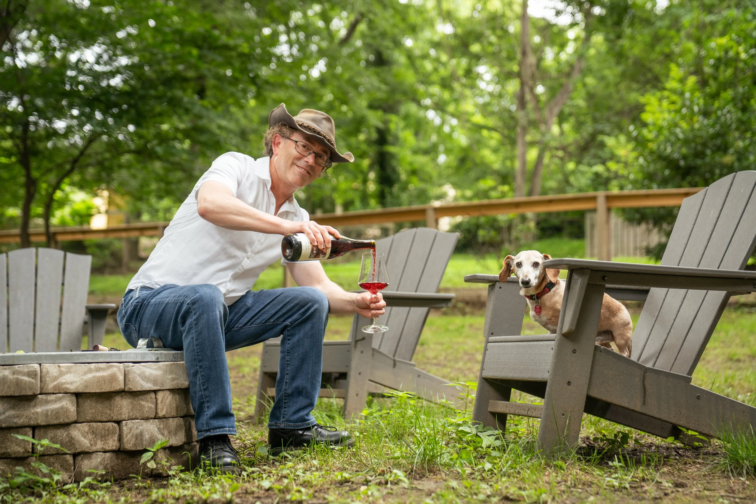 Erik sitting next to an unlit fire pit pouring wine into a glass and his dachshund, Red, sits in a chair across from him.