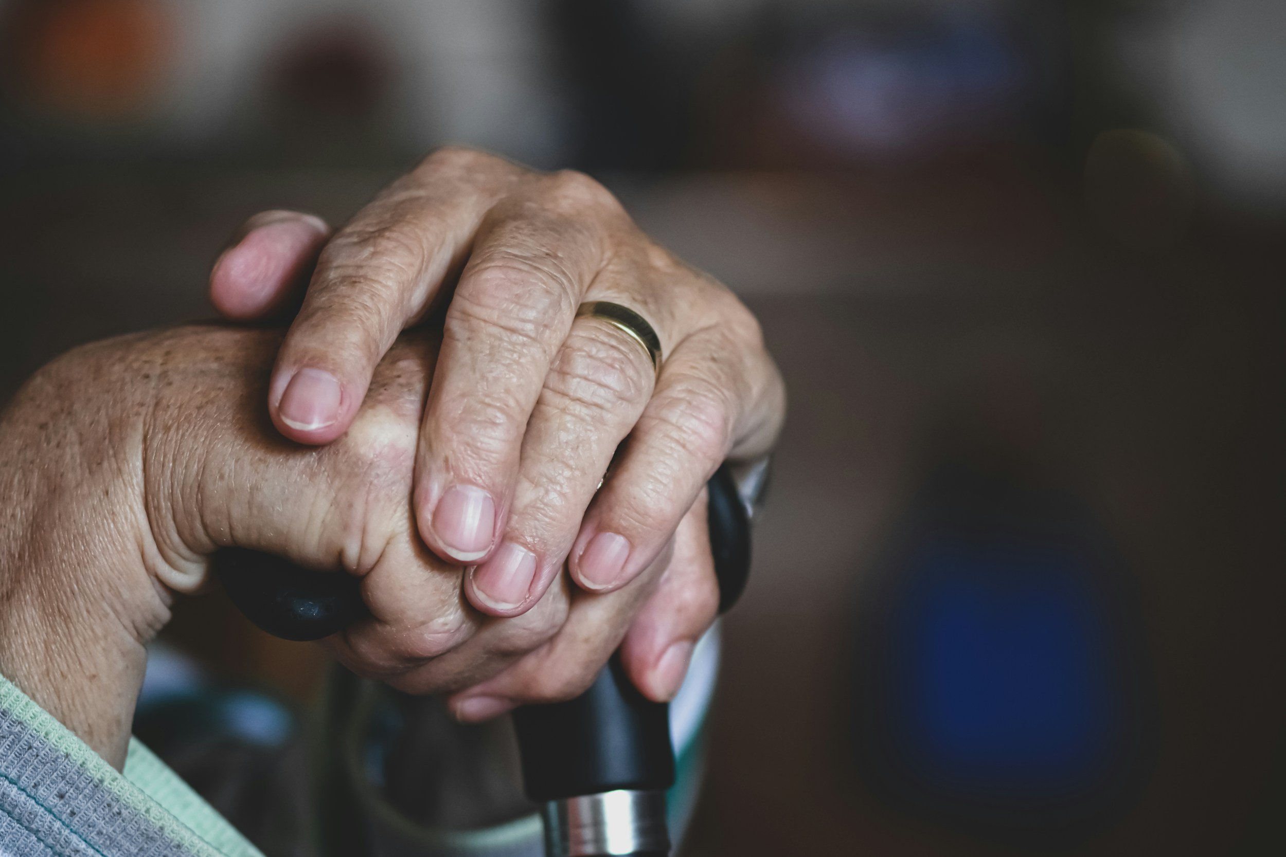 Close-up of elderly hands holding a cane, wearing a wedding band, with a blurred background.