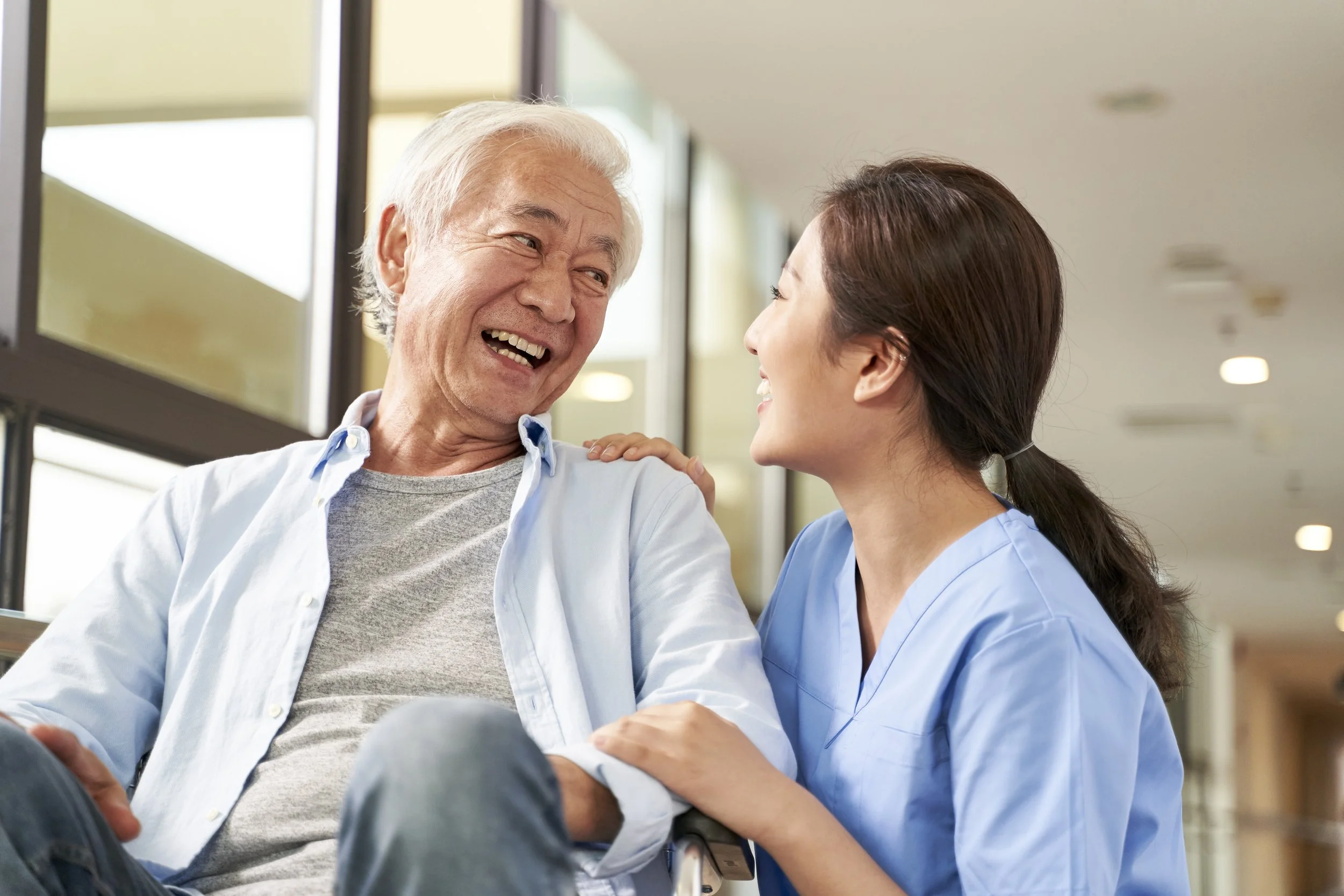 A senior man sitting in a wheelchair, smiling and talking with a young female healthcare worker in blue scrubs, in a hospital corridor.