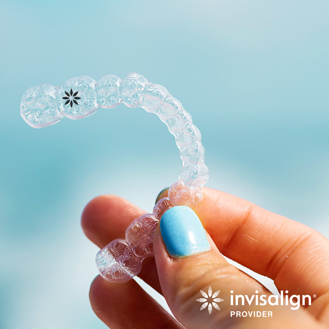 A hand with blue nail polish holding a clear, curved Invisalign aligner against a light blue background.