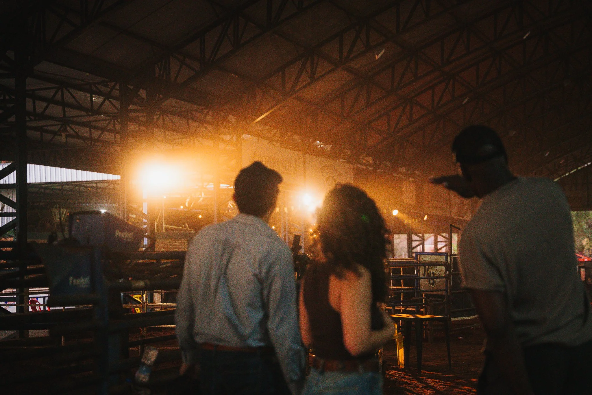 People inside a large industrial-style venue with metal beams, with a bright light source illuminating the background and some individuals visible in the foreground.