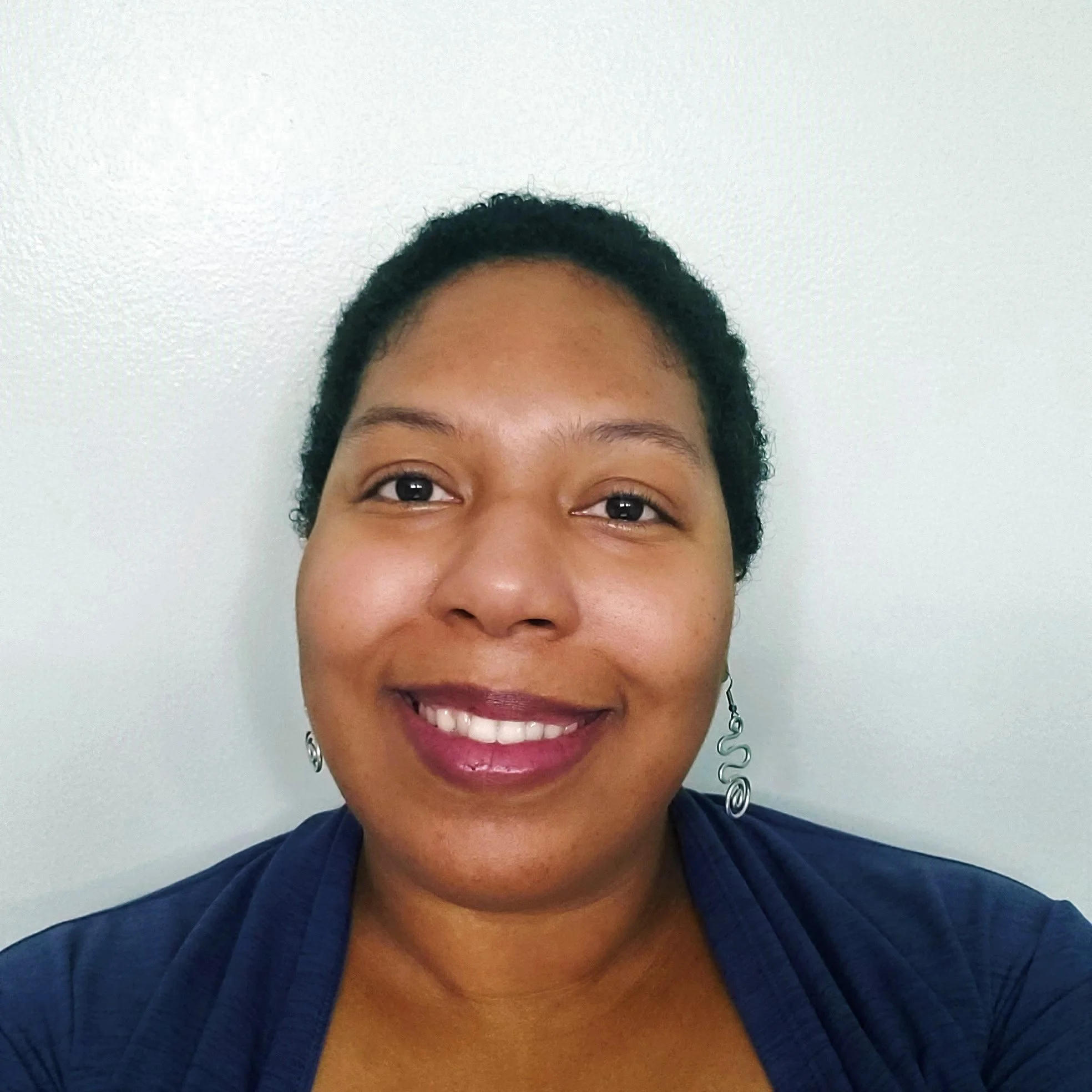 A woman with short black hair, dark skin, and a bright smile, wearing earrings and a dark blue top, against a plain white wall.