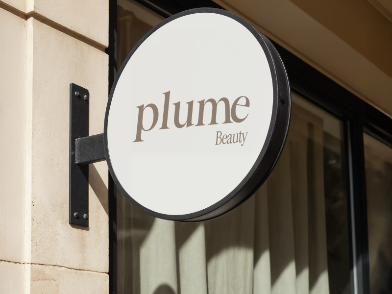 Plume Beauty storefront signage mockup featuring an elegant taupe serif wordmark and Beauty lettering on a circular ivory sign with a black metal frame, mounted on a stone building facade.