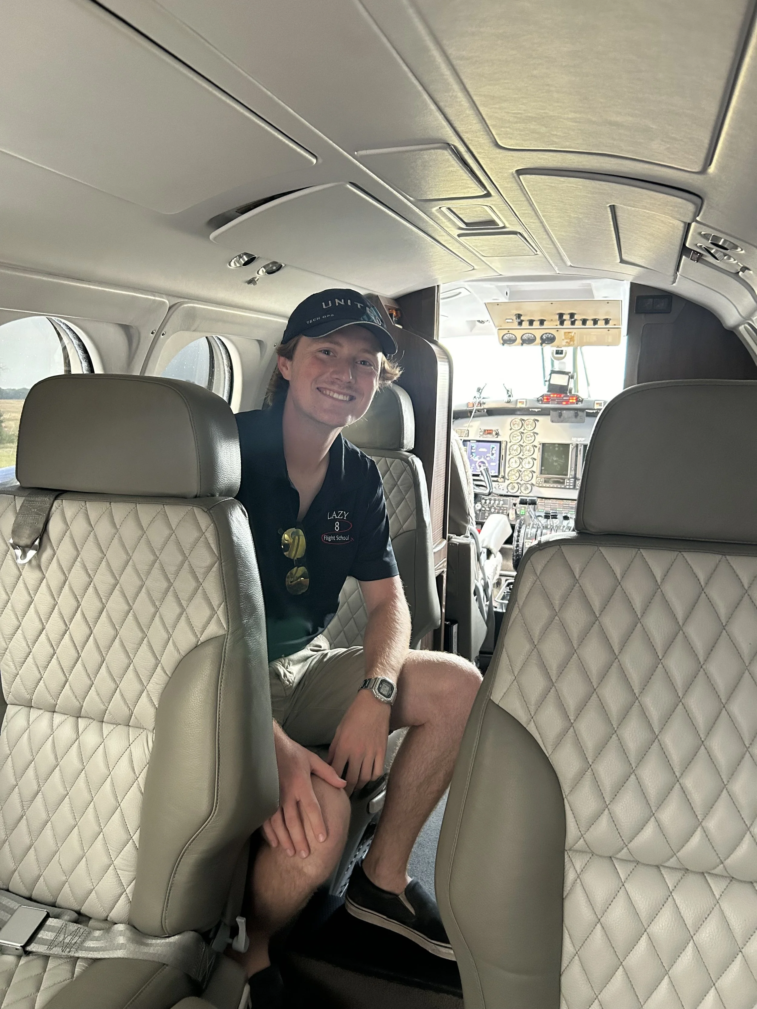 A young man sitting in the cabin of an airplane, smiling and looking at the camera, with the cockpit in the background.