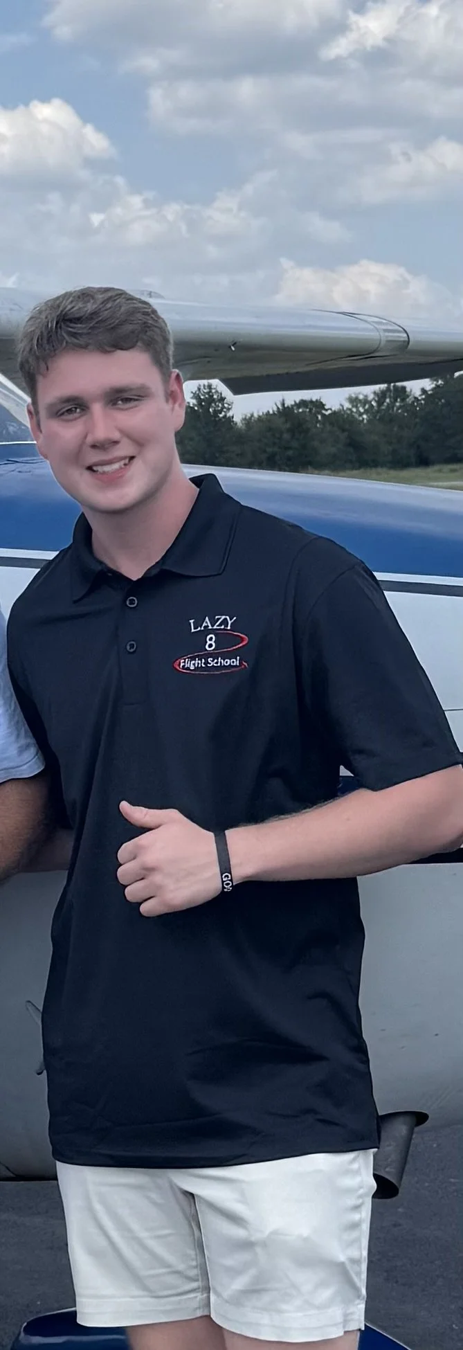 Young man with light brown hair smiling, wearing a black polo shirt with embroidered logo and white shorts, standing outdoors by an airplane.