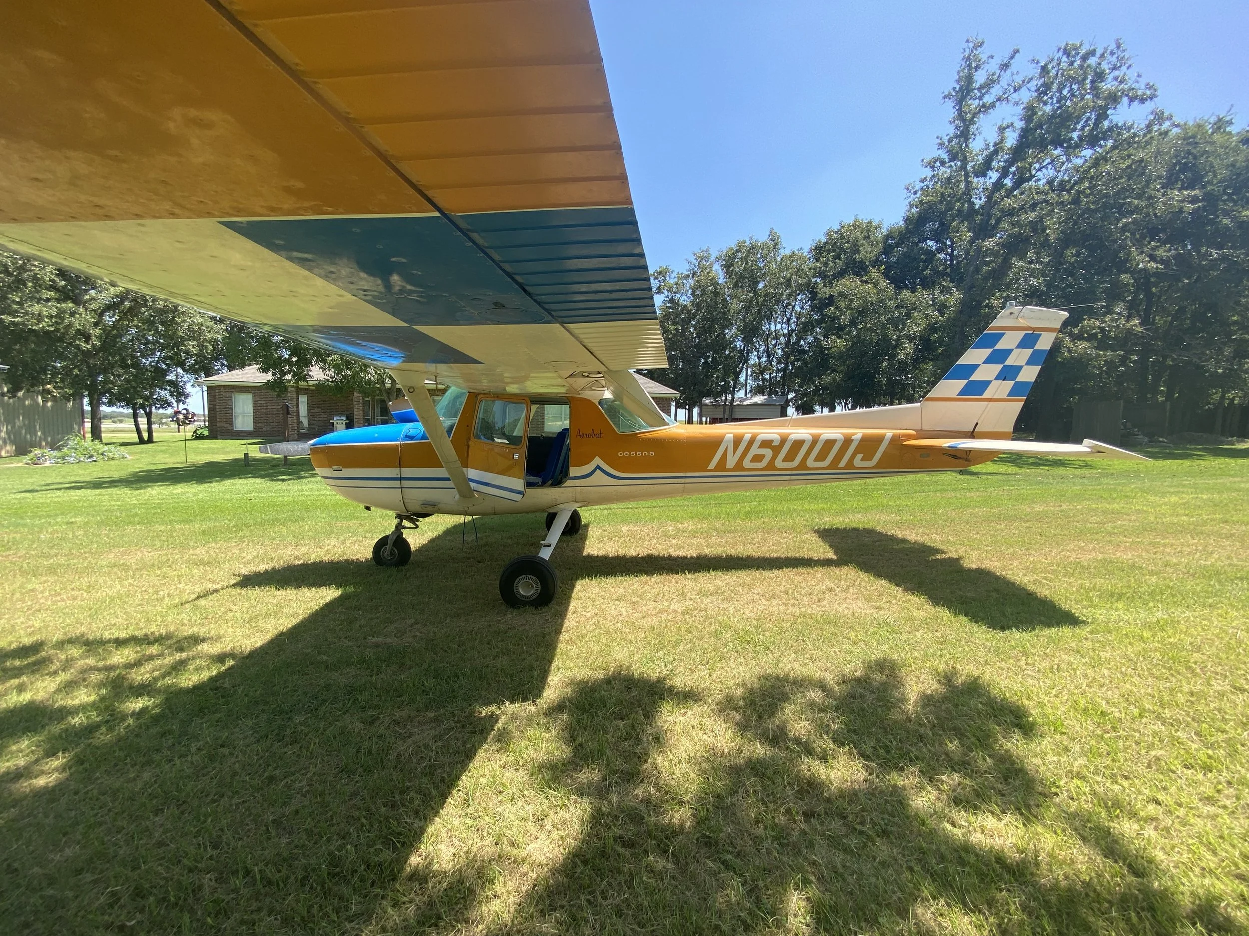 Small single-engine airplane with yellow, blue, and white colors parked on a grassy field, casting a shadow, with trees and a house in the background under a clear blue sky.