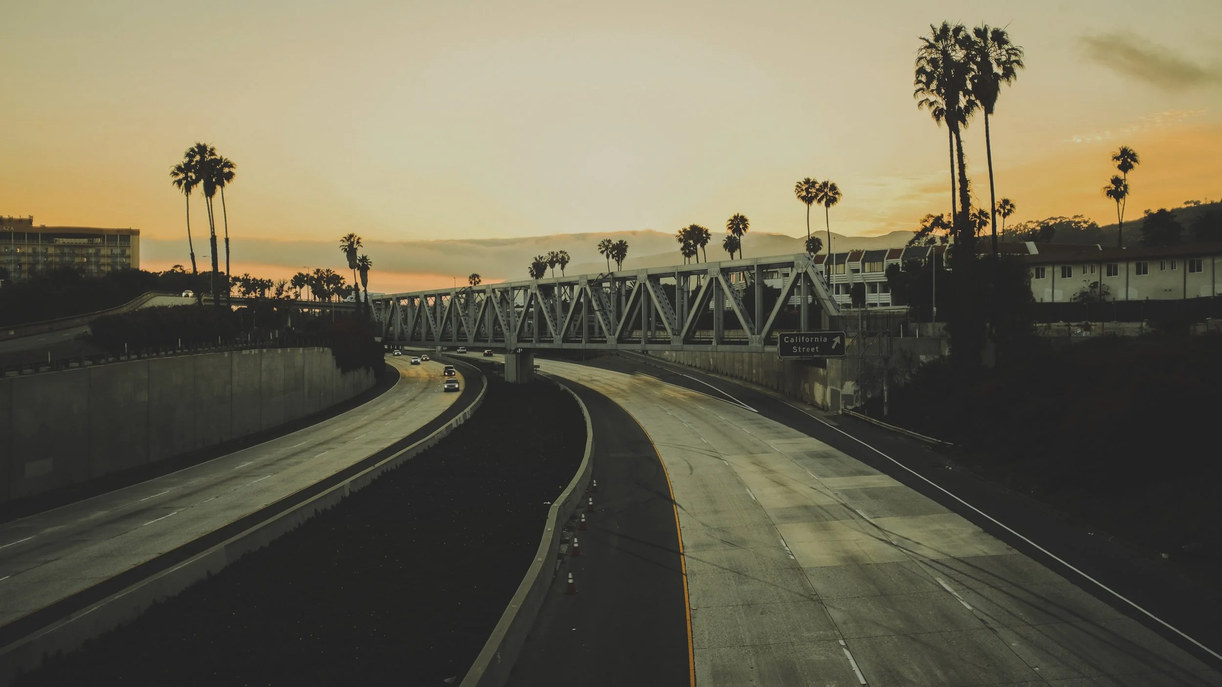 Empty highway with cars, palm trees, and a metal bridge during sunset in California.