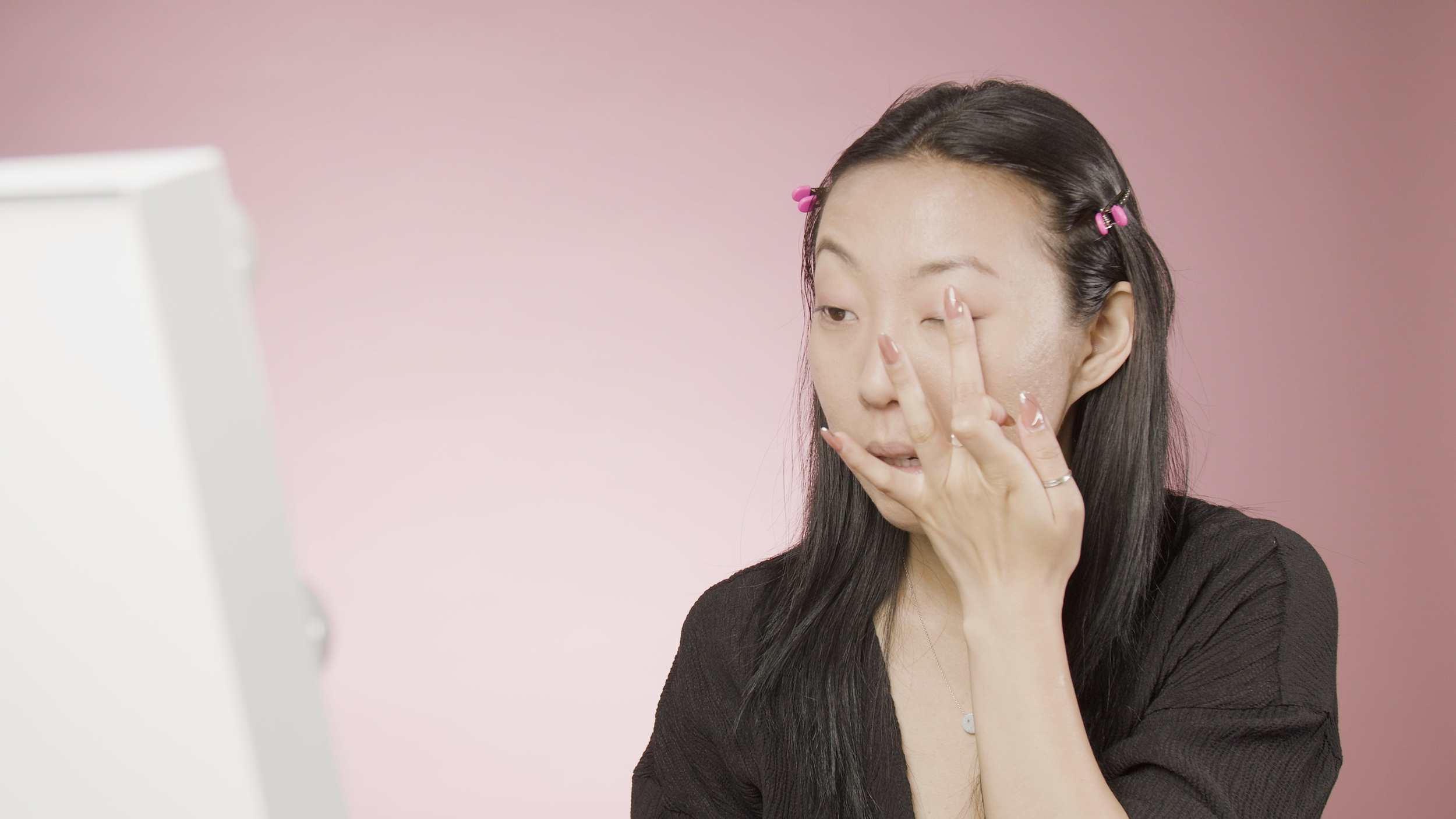 Person applying makeup in front of a mirror with a pink background.