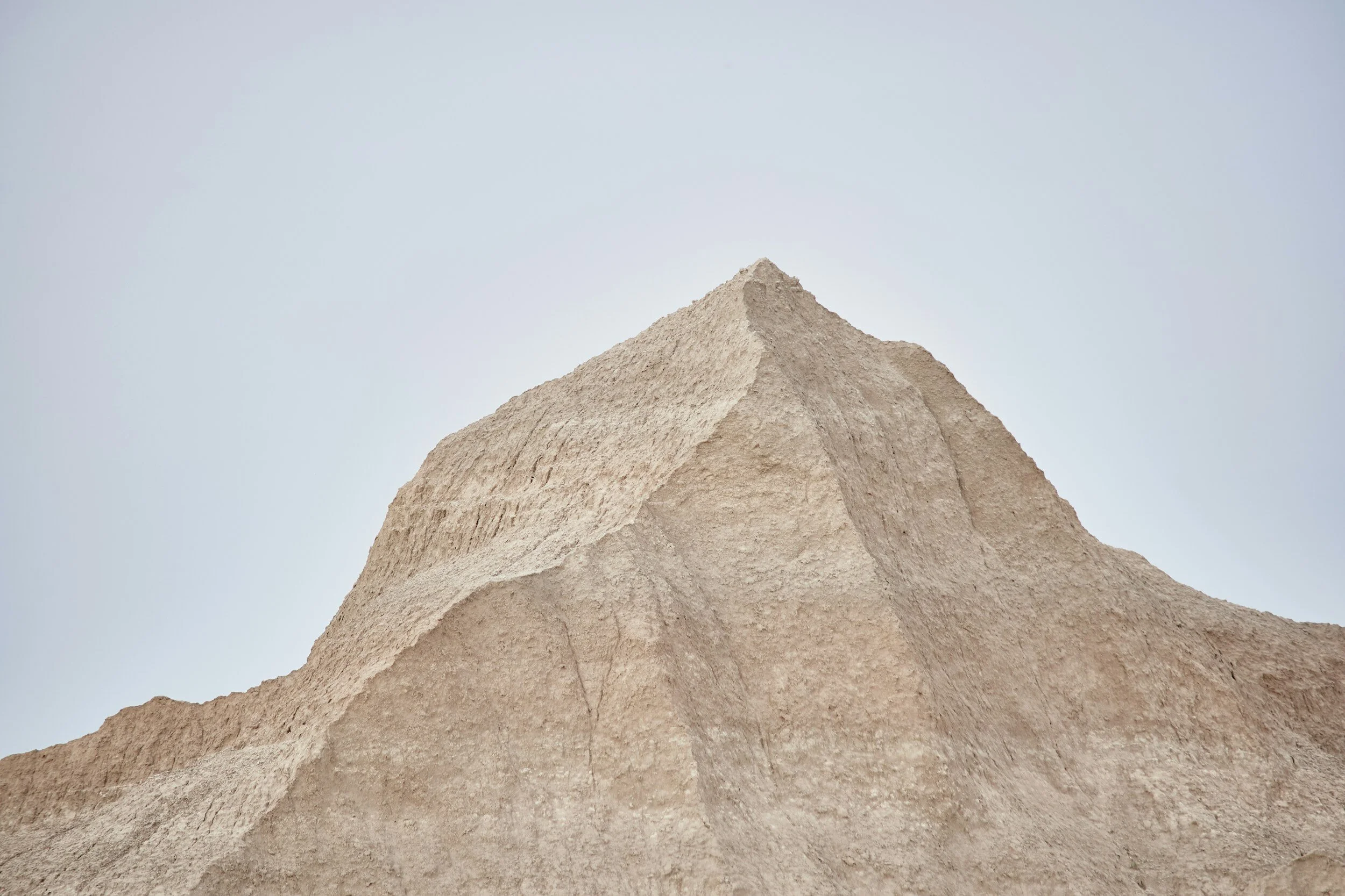 Close-up photo of a large, beige sand or dirt mound against a clear, light blue sky.