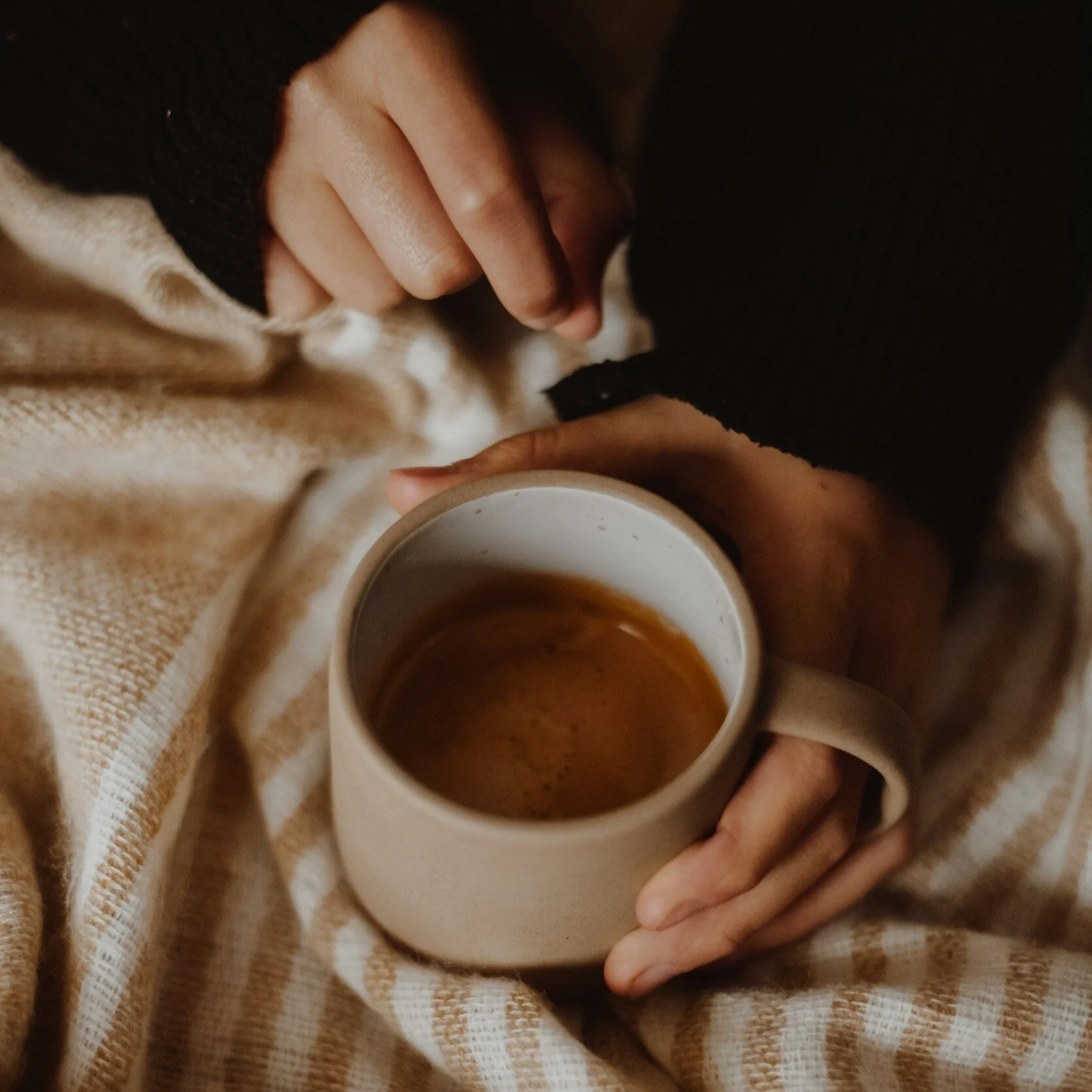 A person holding a beige mug filled with coffee, sitting on a beige and white checked blanket, wearing a black sweater and cream cardigan.
