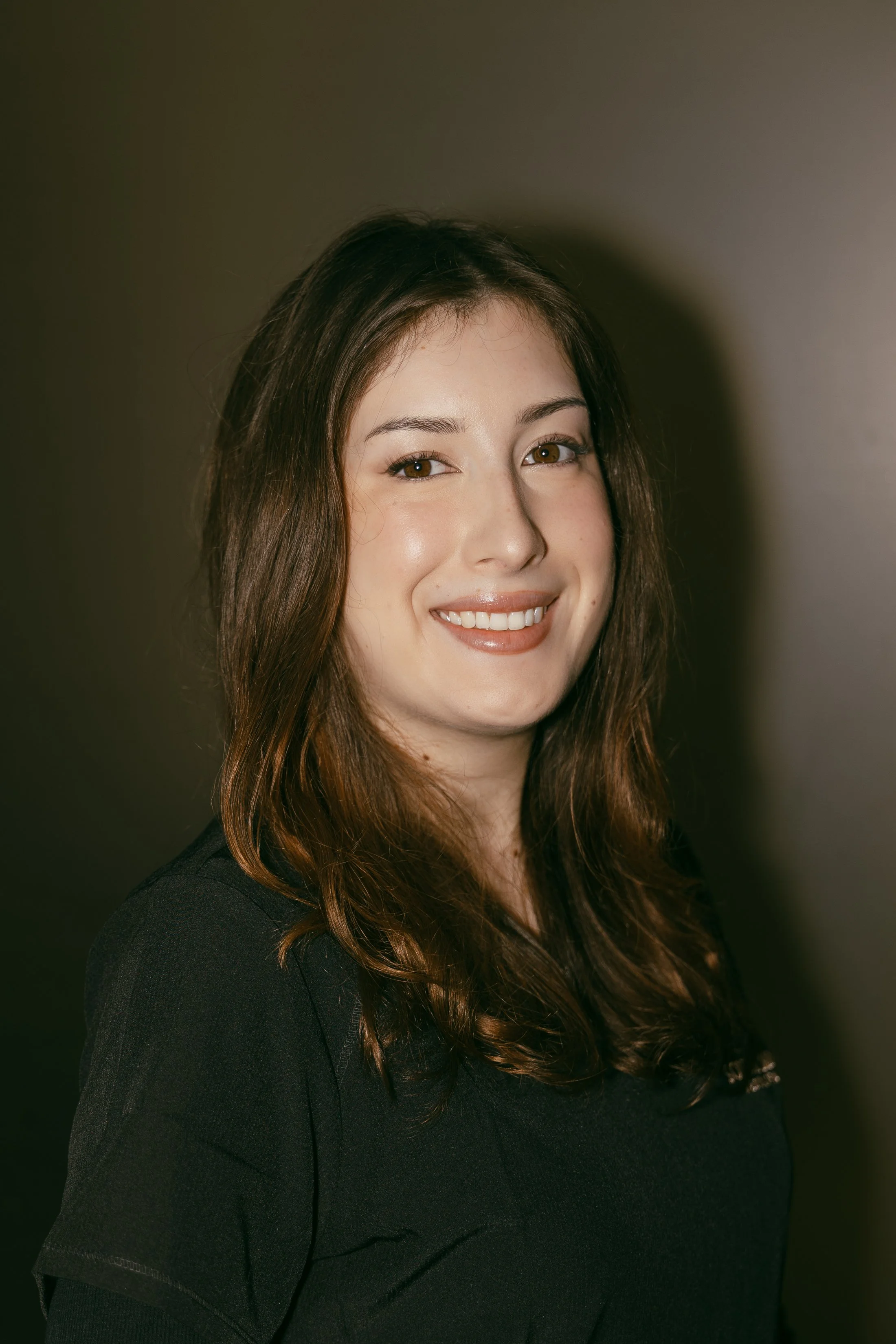 A woman with long brown hair smiling, wearing a black top, against a plain dark background.