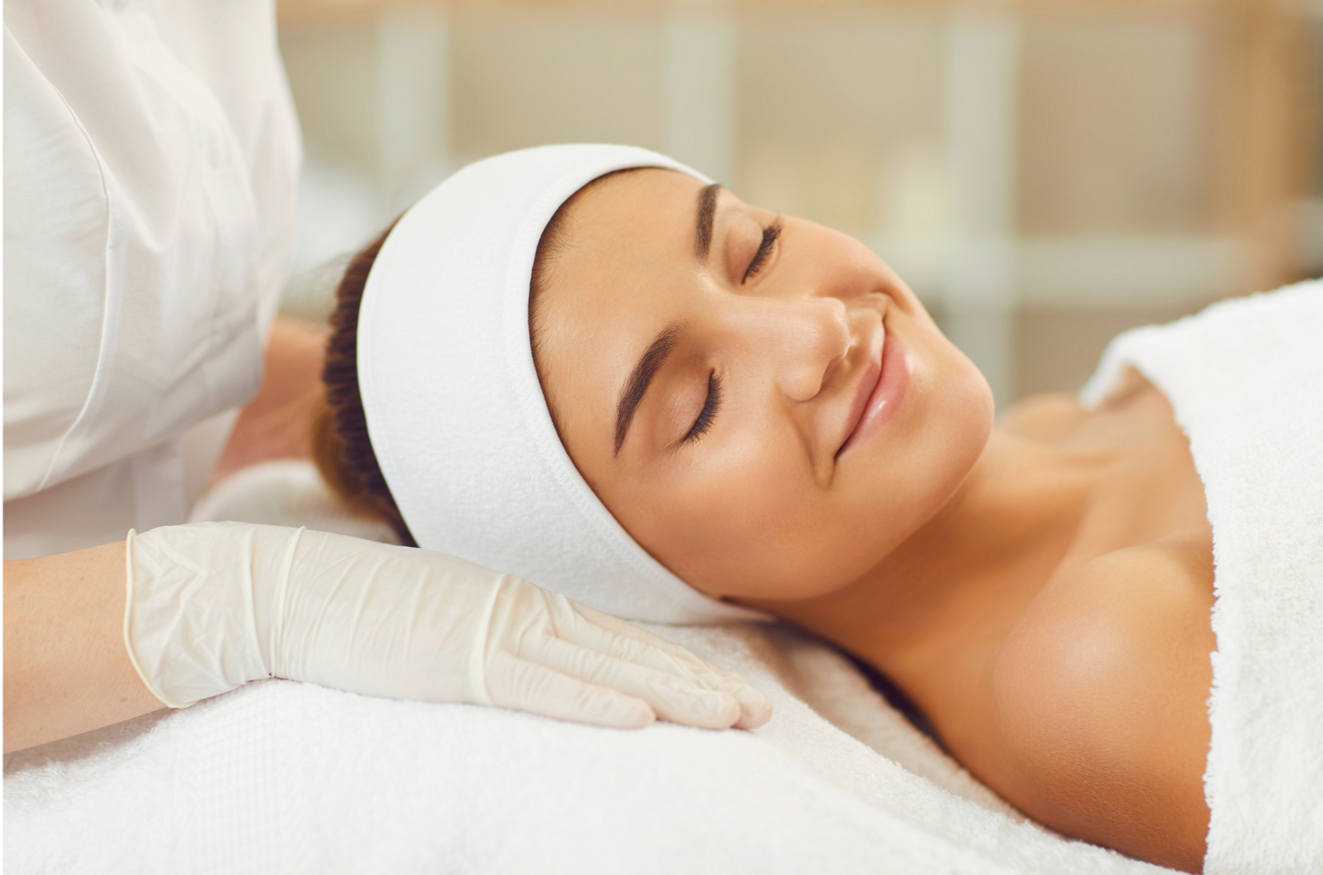Woman receiving a facial treatment at a spa, lying on her back with eyes closed, wearing a white headband, while an esthetician applies a treatment with gloved hands.