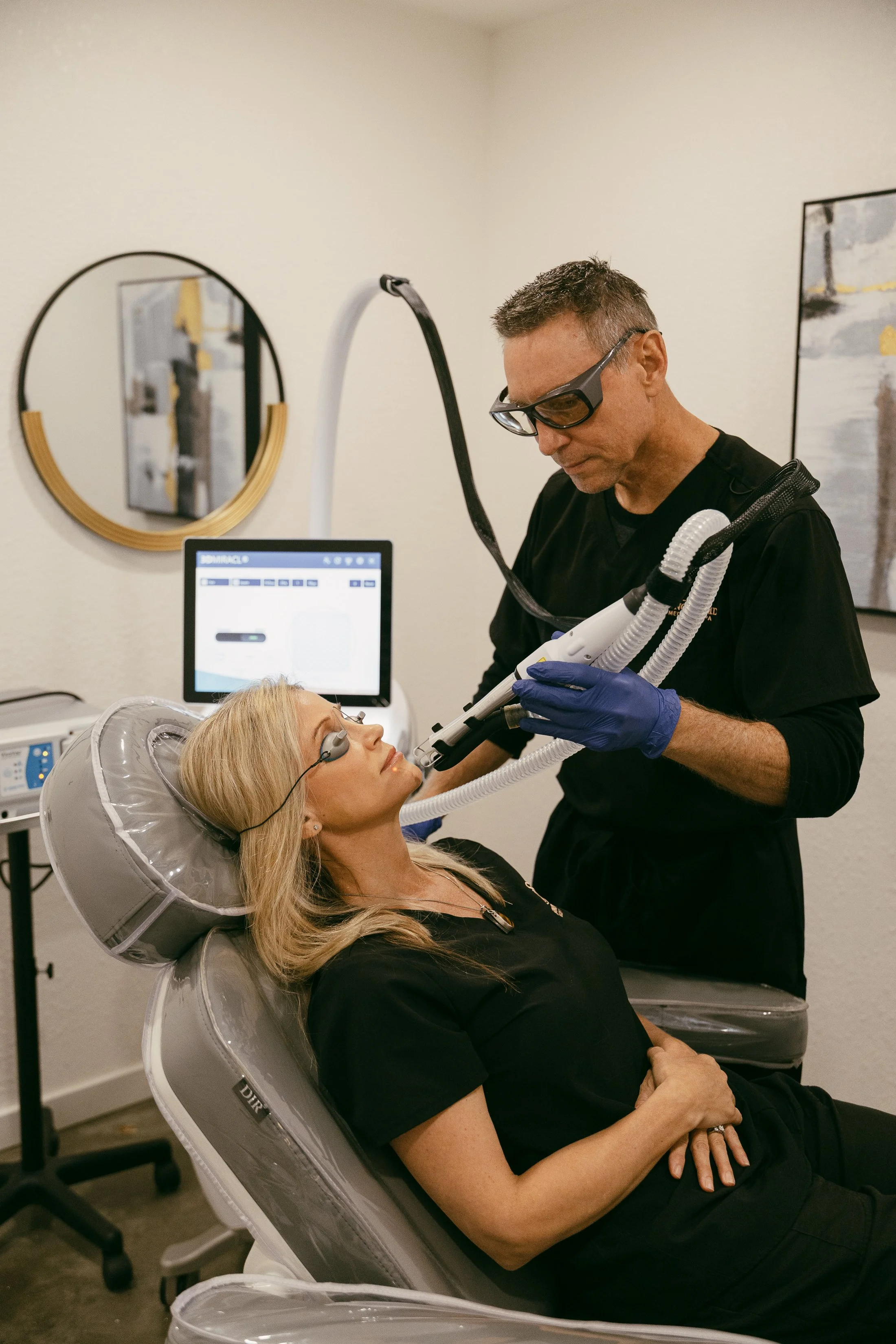 A woman lying in a dental or medical chair receiving a cosmetic or dermatological treatment from a professional, who is using specialized equipment.