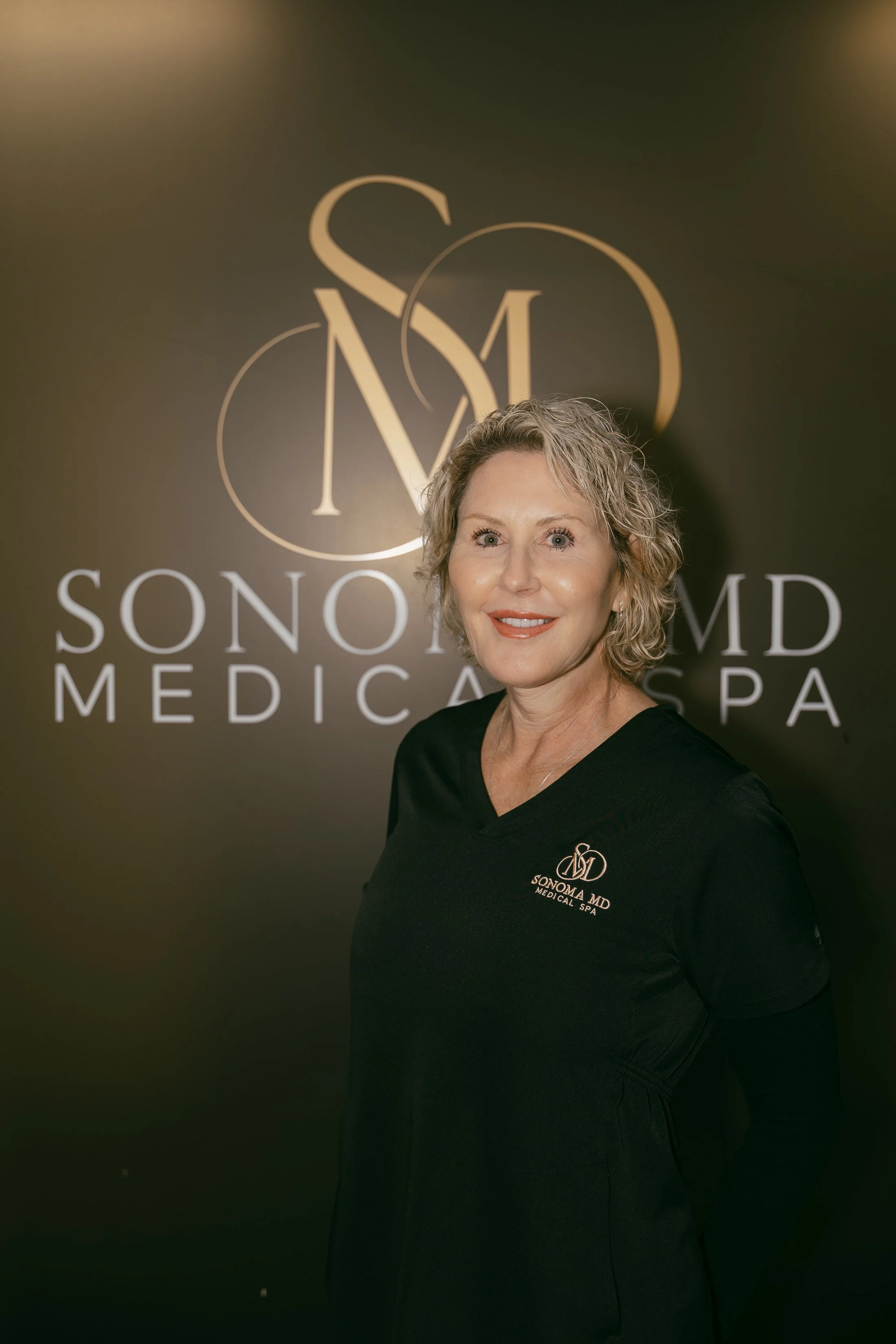 A woman with short curly gray hair smiling in front of a Sonoma MD Medical Spa logo backdrop.