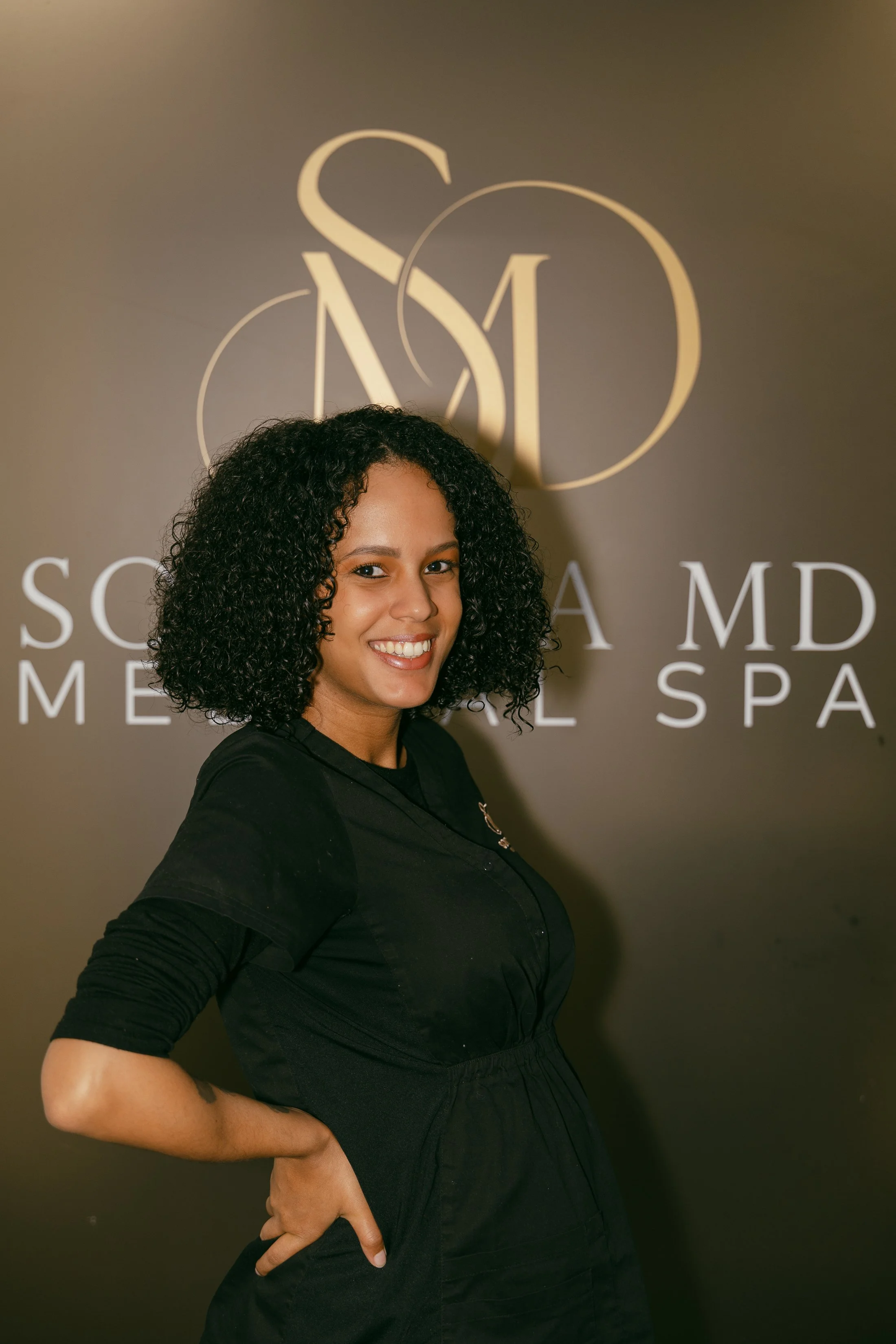 Woman with curly hair smiling, standing in front of a gray wall with the logo of Sola MD Medical Spa.