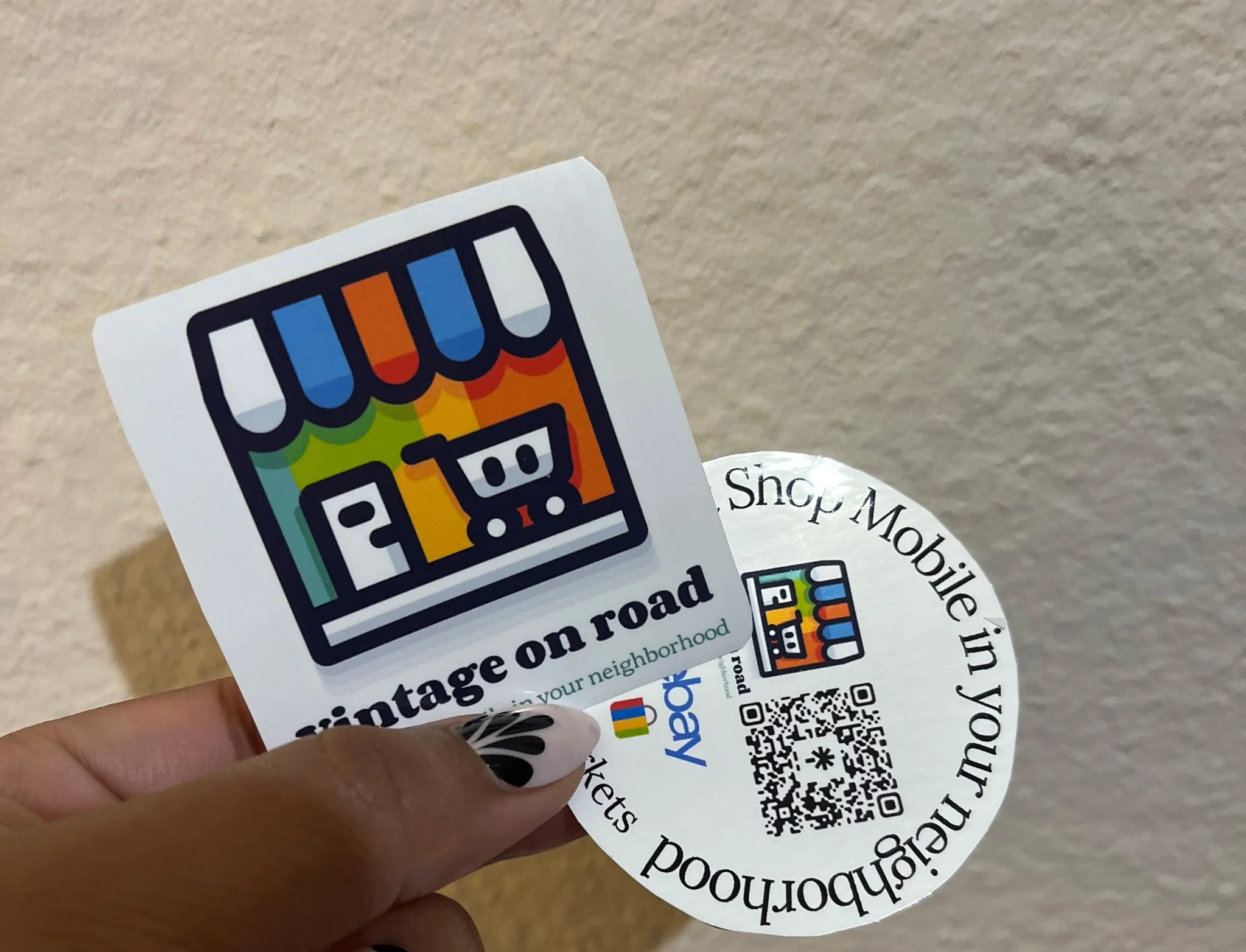 Person holding printed stickers or labels with a colorful logo of Vintage on road and a QR code, with a beige wall background and partially visible circular paper.