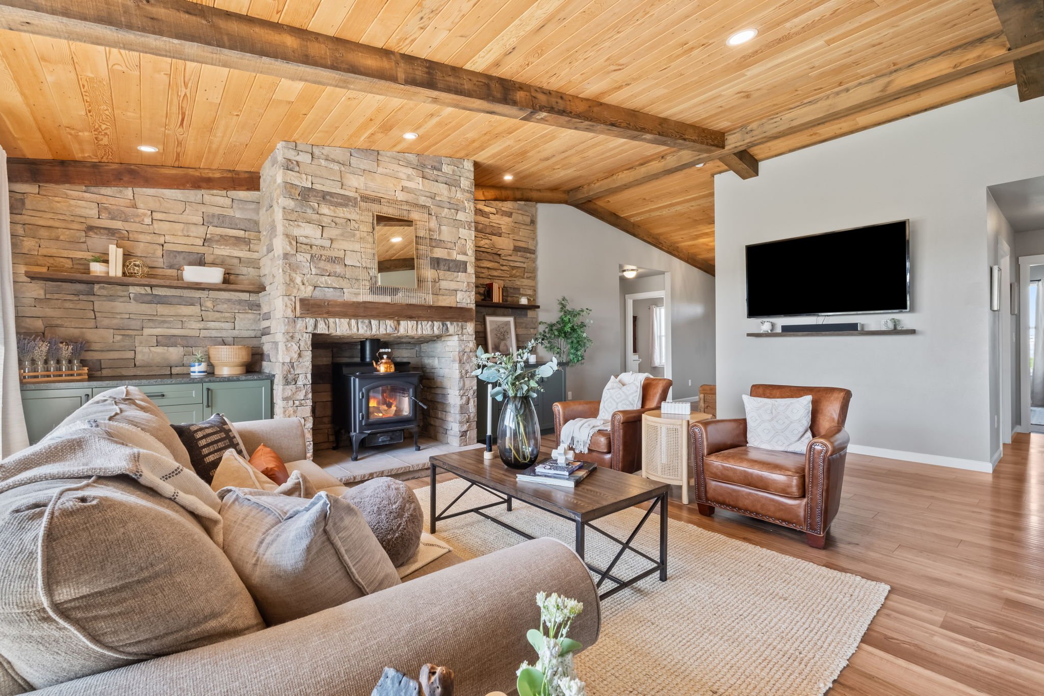 Living room with a stone fireplace, wooden ceiling, and comfortable furniture including a beige sofa and two brown leather armchairs, decorated with pillows and a coffee table with a vase of flowers, in a cozy and modern style.