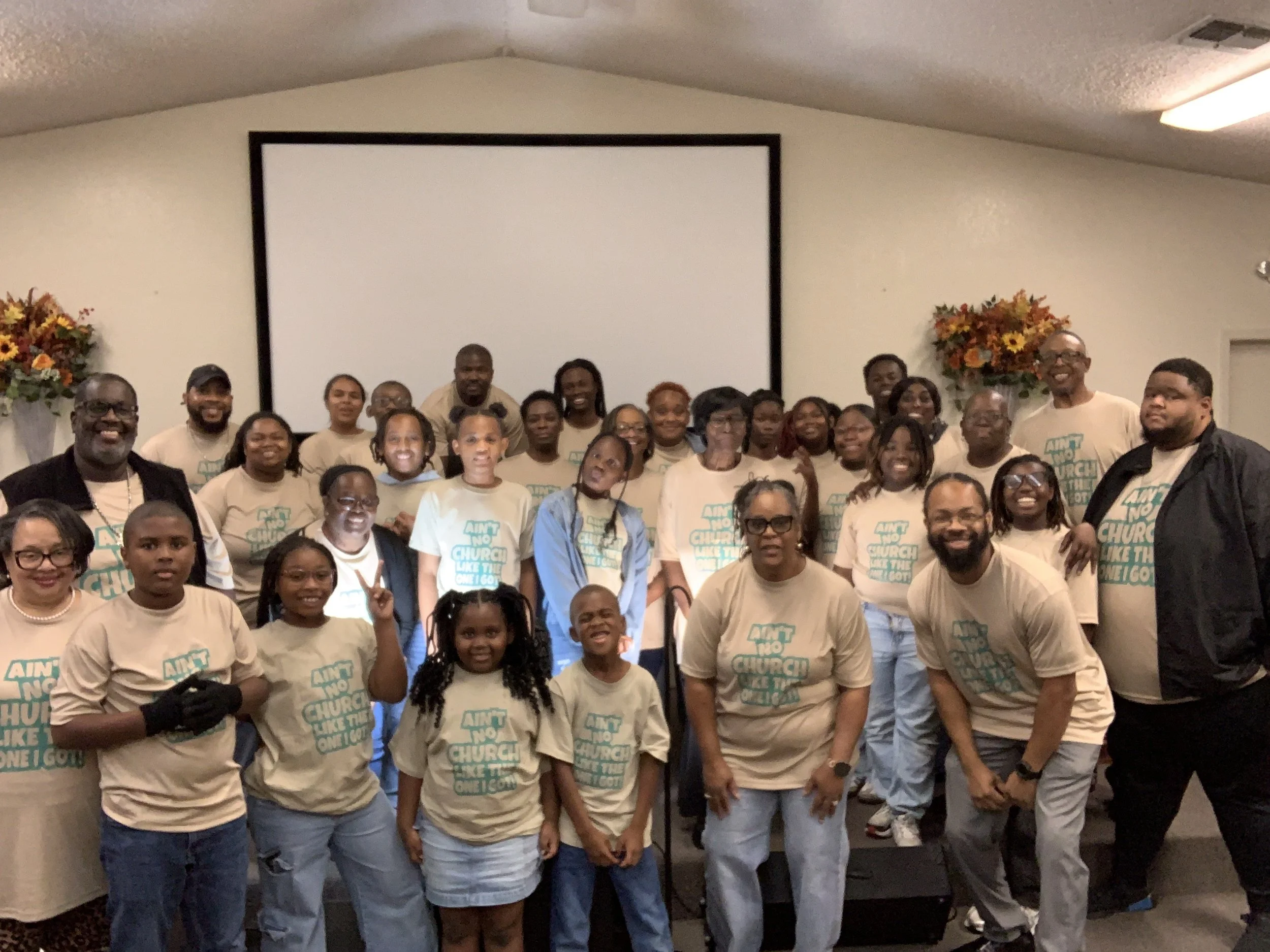 Group of people posing together in a room with a large blank white screen behind them, some wearing matching beige T-shirts with teal text.