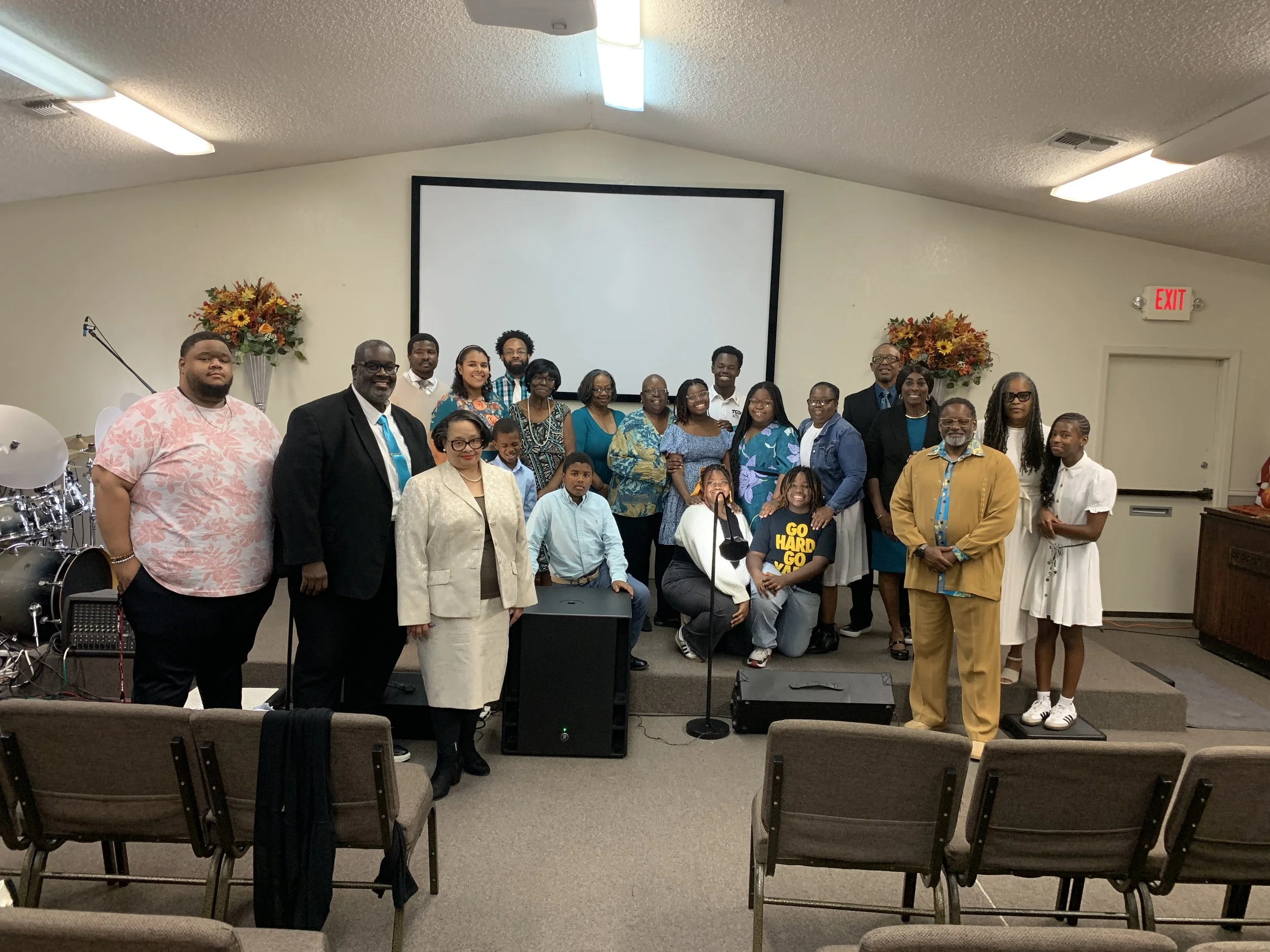 A group of people standing together on a stage in a church or community hall, posing for a photo. There are adults and children, dressed in a mix of formal and casual attire, with a church setting including flowers and musical instruments in the back