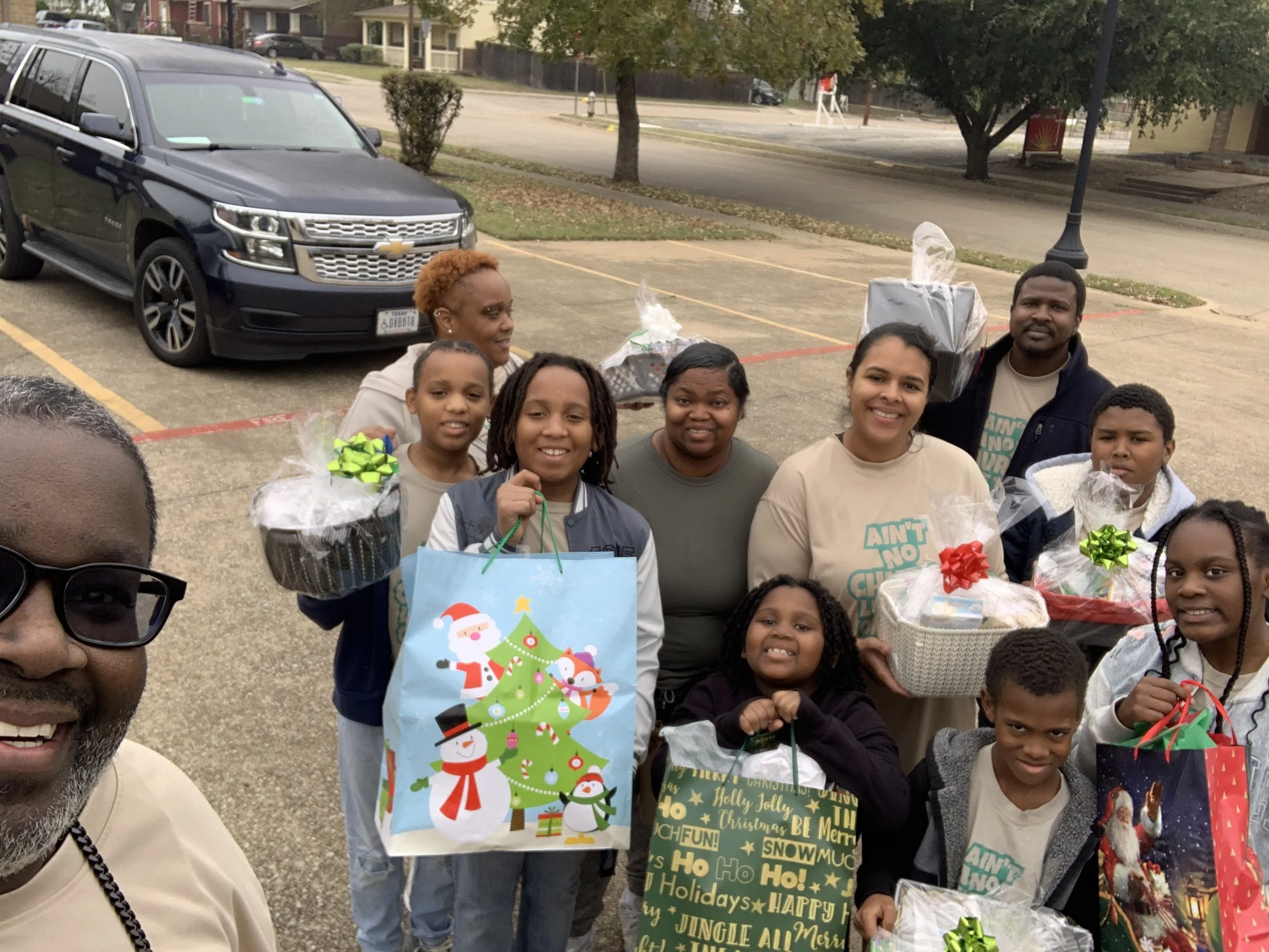 A group of people, including adults and children, are standing in a parking lot decorated with Christmas gifts, some with bows, and Christmas-themed bags, smiling at the camera. There are cars and trees in the background.