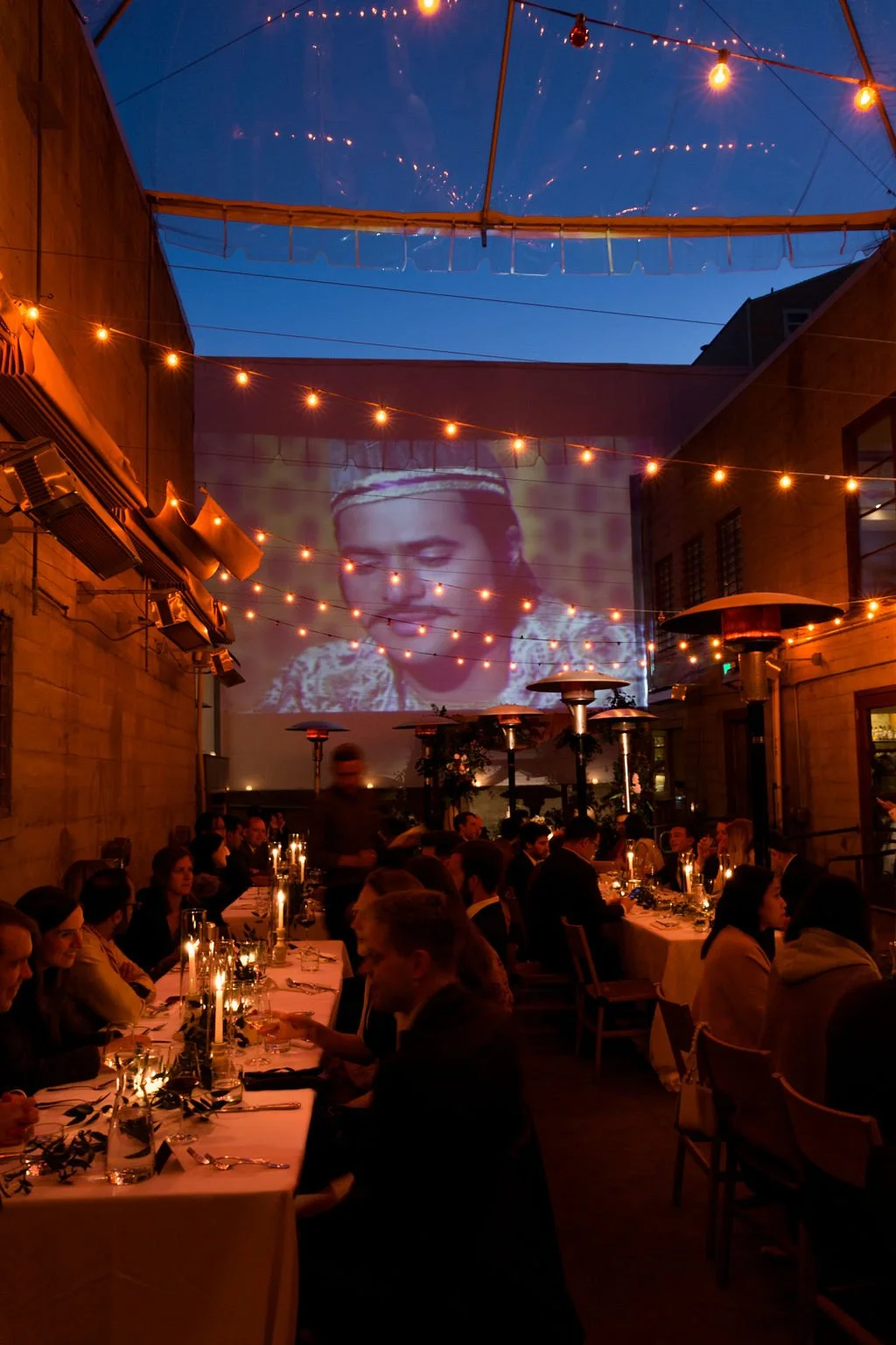 A nighttime outdoor dinner party with string lights, patio heaters, and a large projection of a woman's face on the wall. Guests are seated at long tables with candlelit centerpieces.