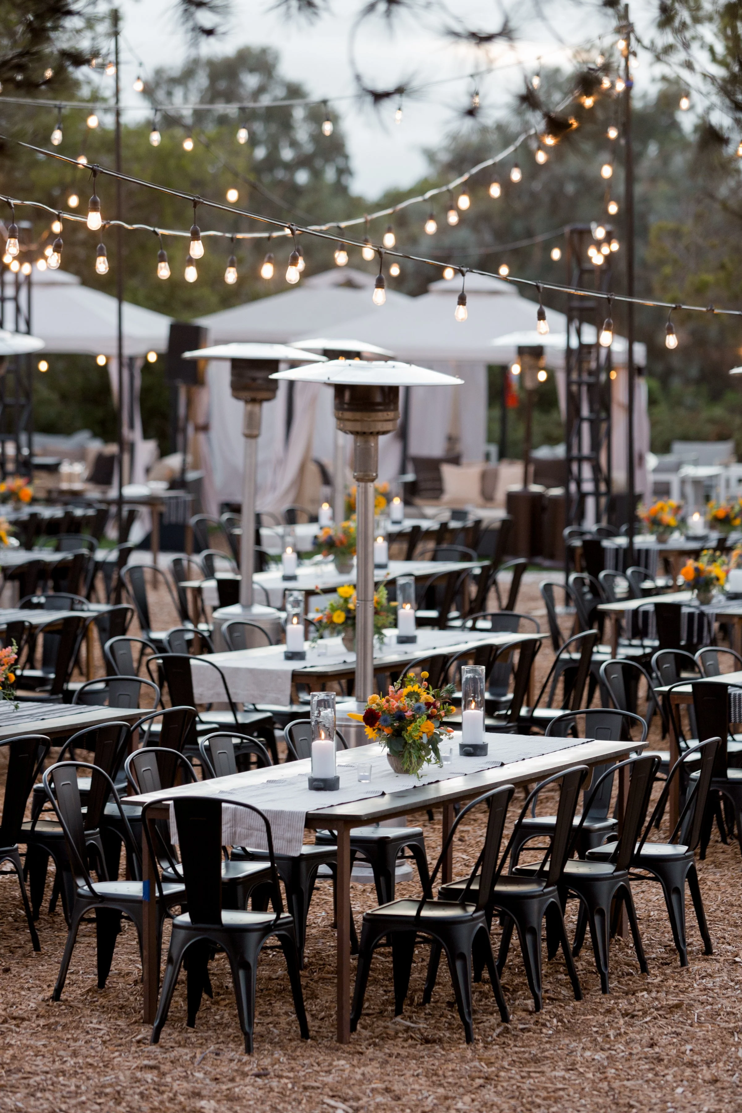 Outdoor event setup with tables decorated with flower arrangements and lit candles, string lights overhead, and patio heaters, likely for a dinner or celebration.