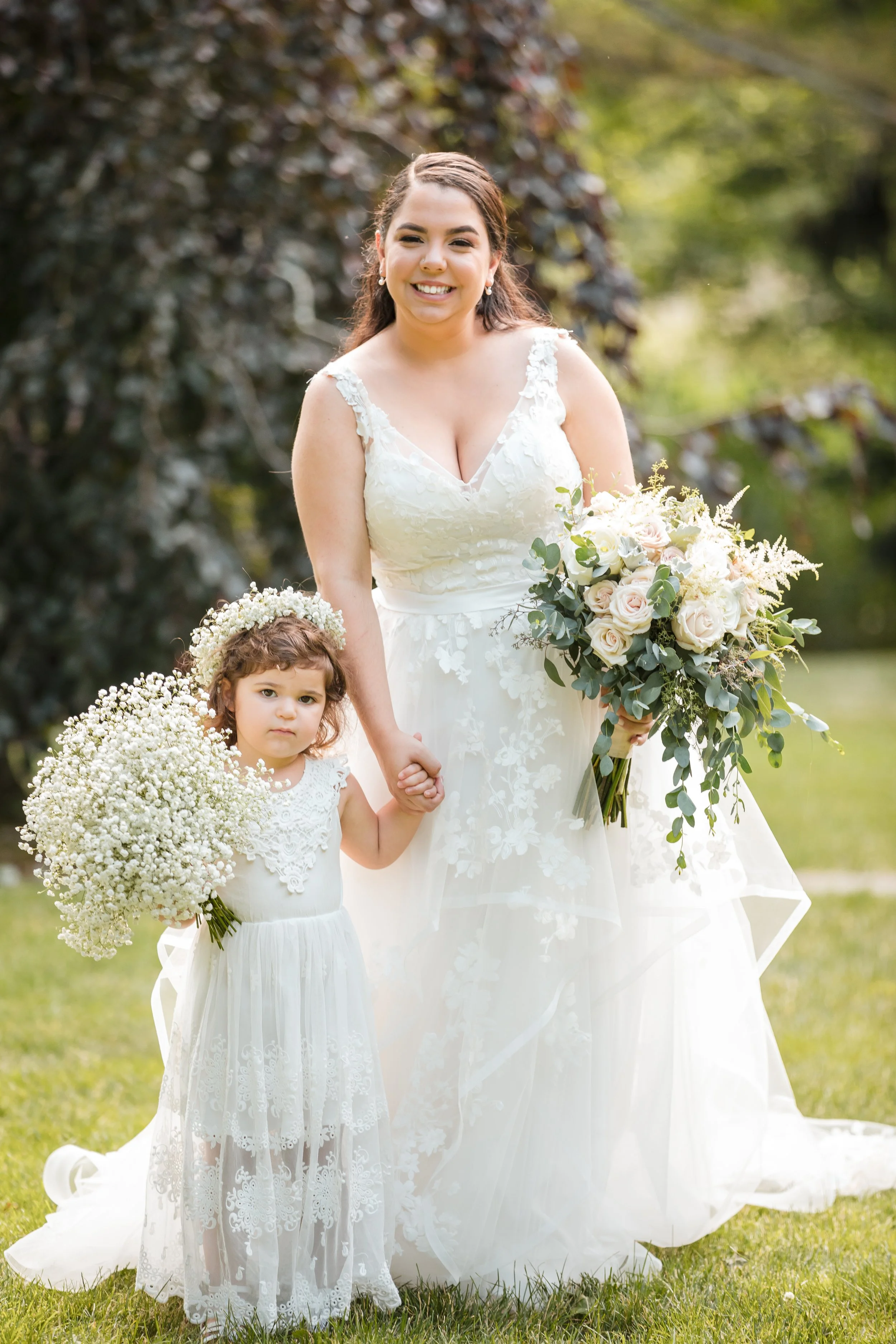 A woman in a white wedding dress holding a bouquet of flowers and a young girl in a white dress holding a large bouquet of baby’s breath flowers, both standing outdoors on grass with trees in the background.