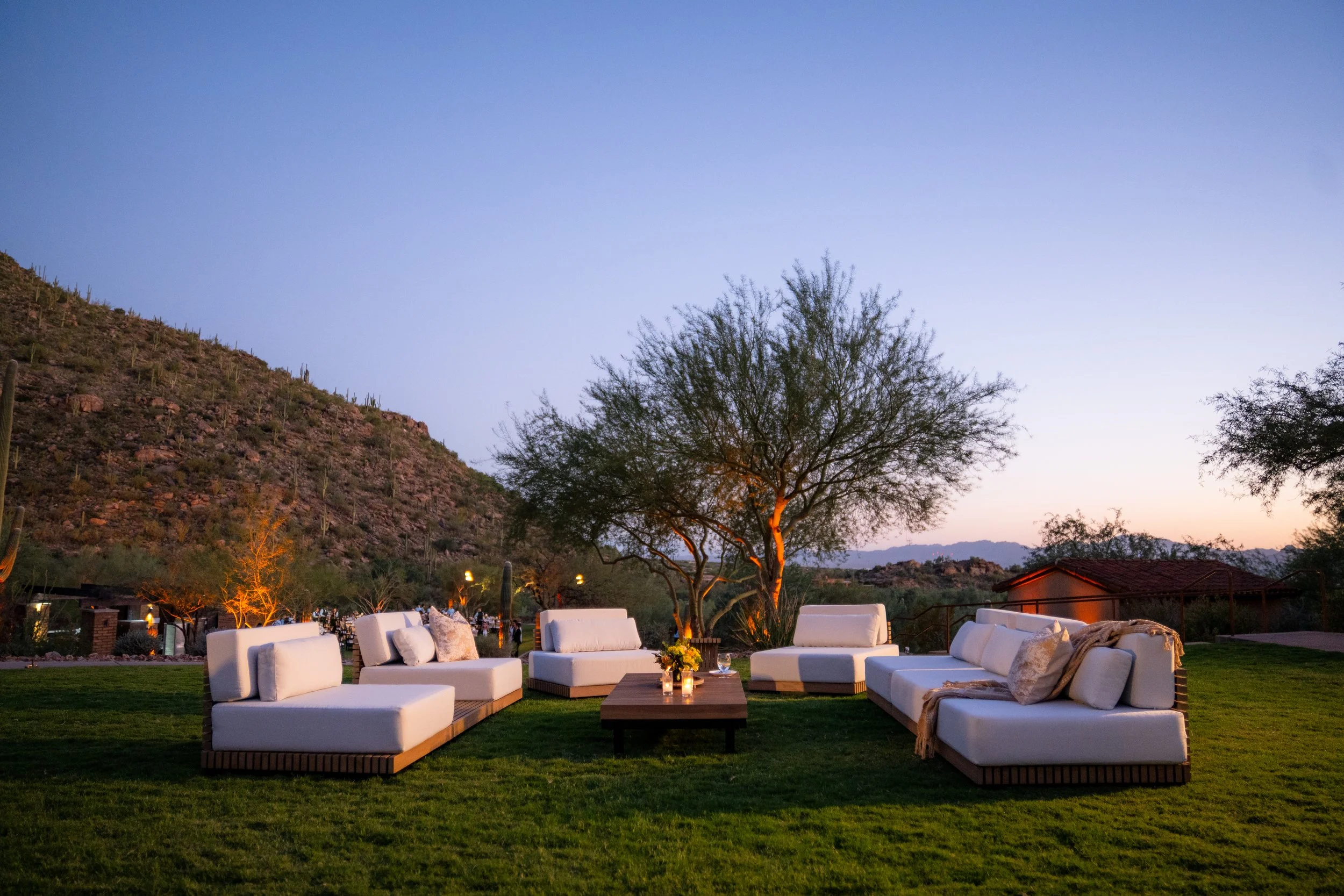Outdoor living area with white cushioned sofas arranged in a semi-circle around a wooden coffee table, with a floral arrangement and candles on it, set on a grassy lawn during evening with mountain and desert landscape in the background.