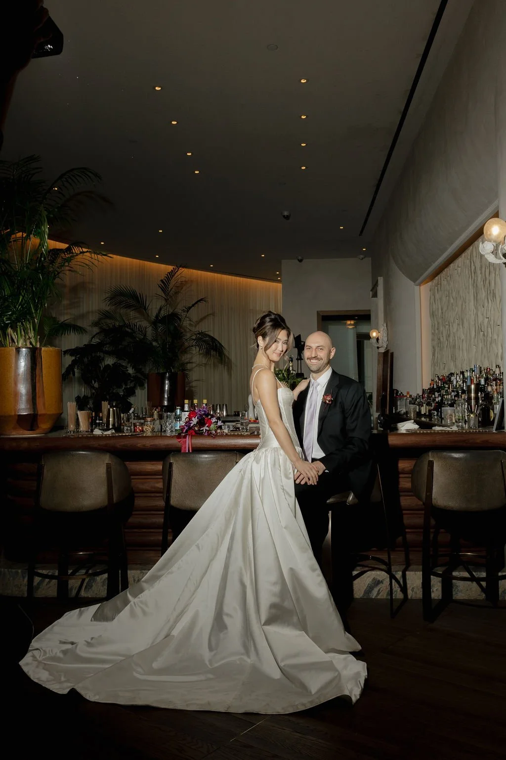 A newlywed couple is standing in a bar, holding hands and smiling at the camera. The bride is wearing a white wedding gown with a long train, and the groom is dressed in a black suit. The bar area has barstools, plants, and various bottles and glasse