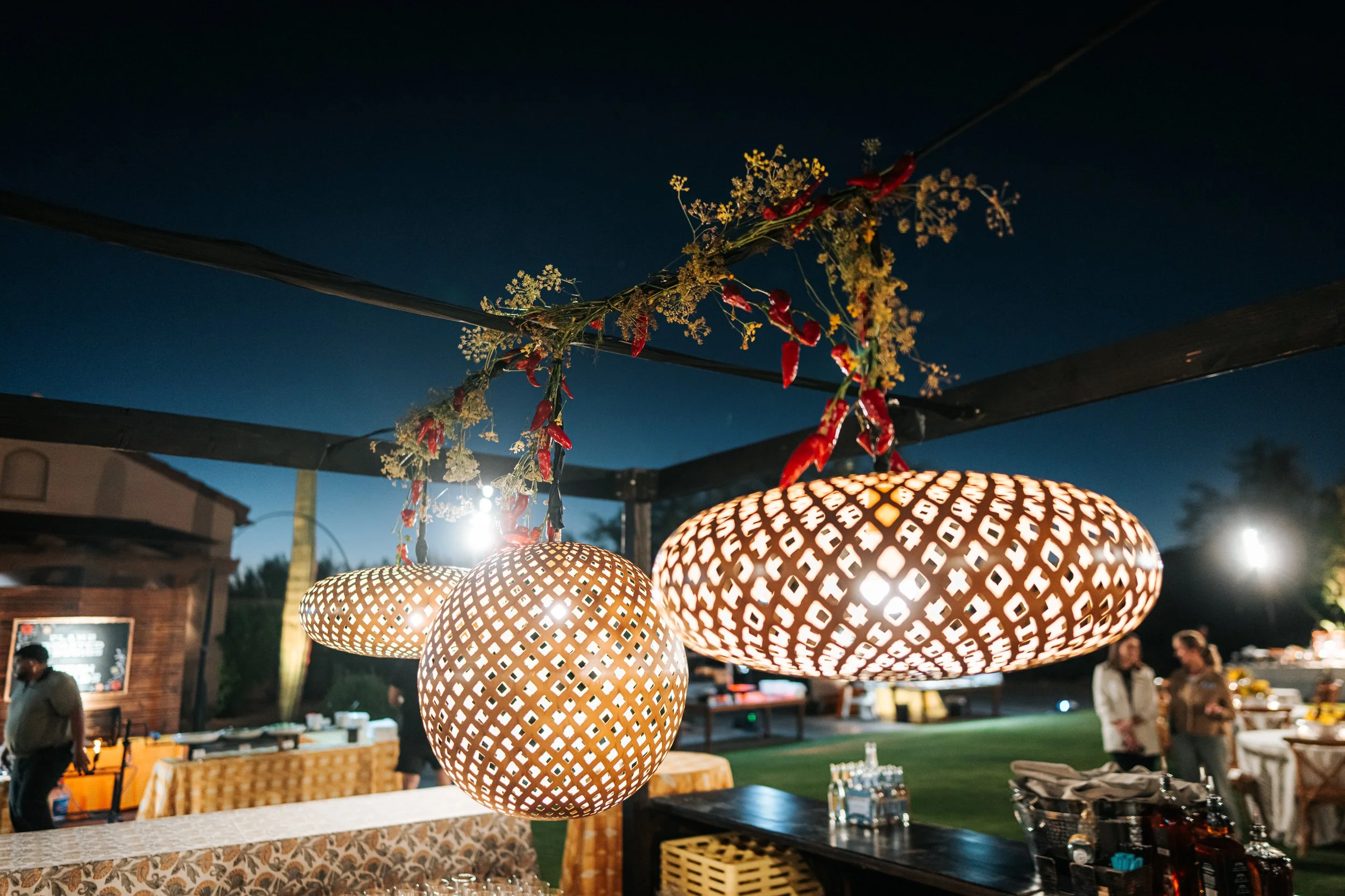 Decorative hanging lanterns and string lights illuminate an outdoor evening event with tables, people, and a chalkboard menu.