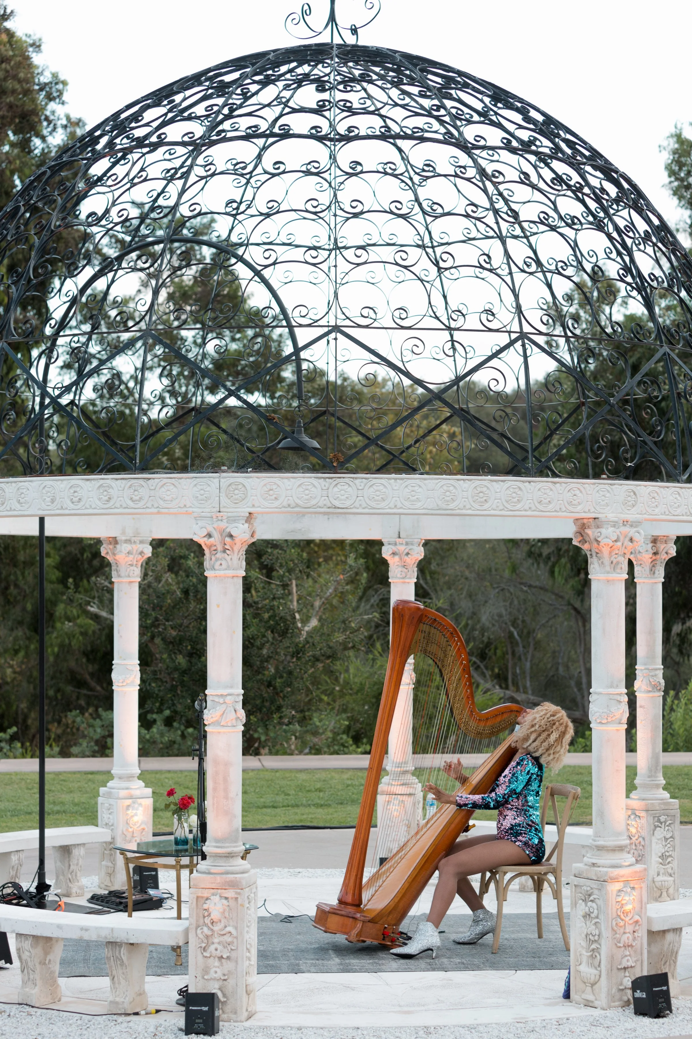 A woman with curly blonde hair playing an acoustic harp inside a white marble gazebo with intricate columns and a domed iron lattice ceiling, set in an outdoor garden with trees in the background.