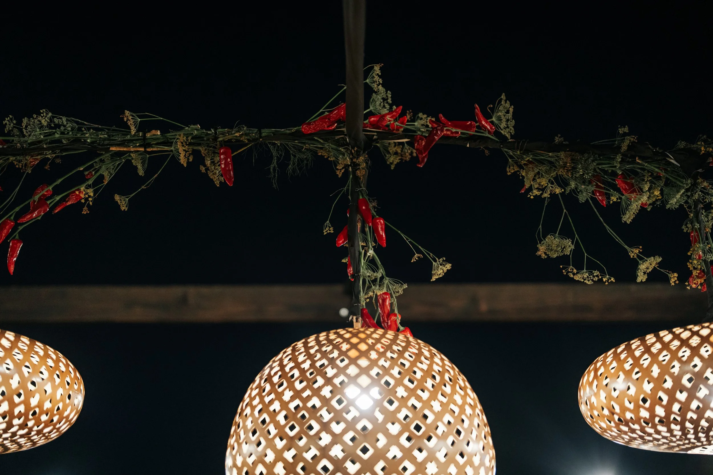 Hanging decorative lampshades with intricate cutout patterns, illuminated in a dark setting, with a garland of flowers and red chili peppers above them.