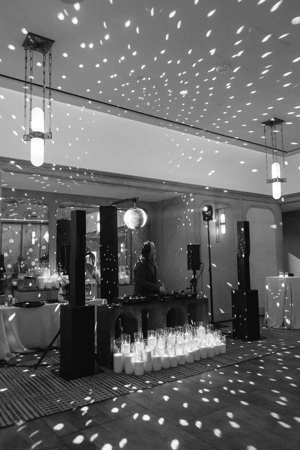 Black and white photo of an indoor event with a DJ playing music, surrounded by candles. The ceiling has a light projection casting patterns across the room, creating a festive atmosphere.