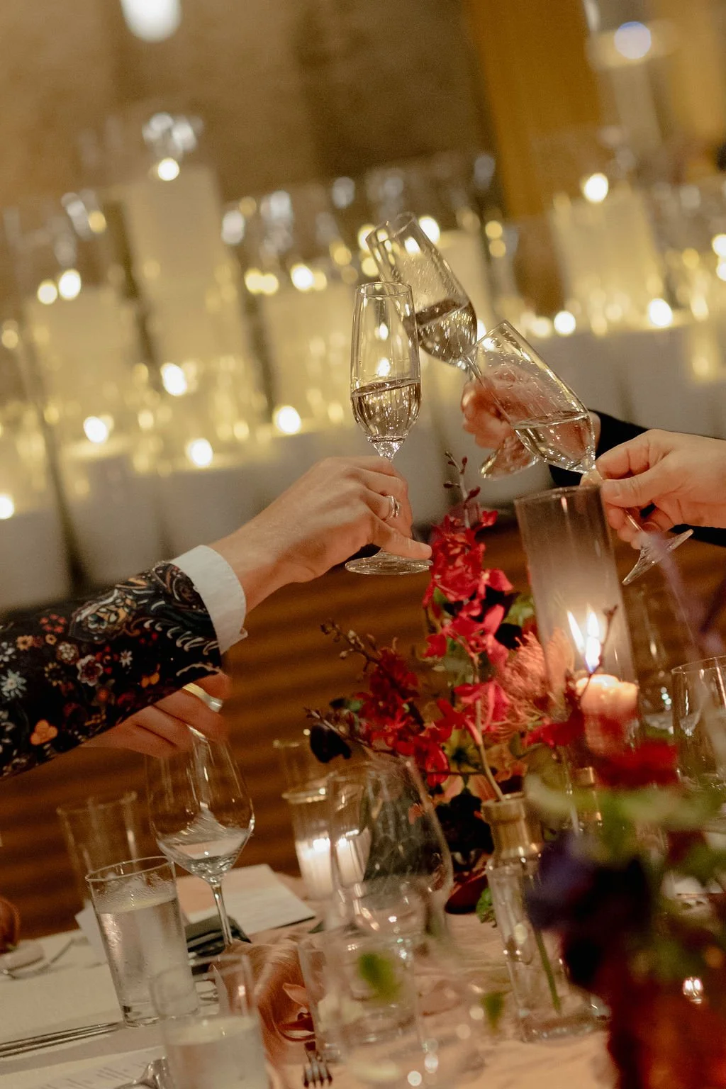 People raising glasses of champagne for a toast at a celebratory dinner with a floral centerpiece and candles on the table.