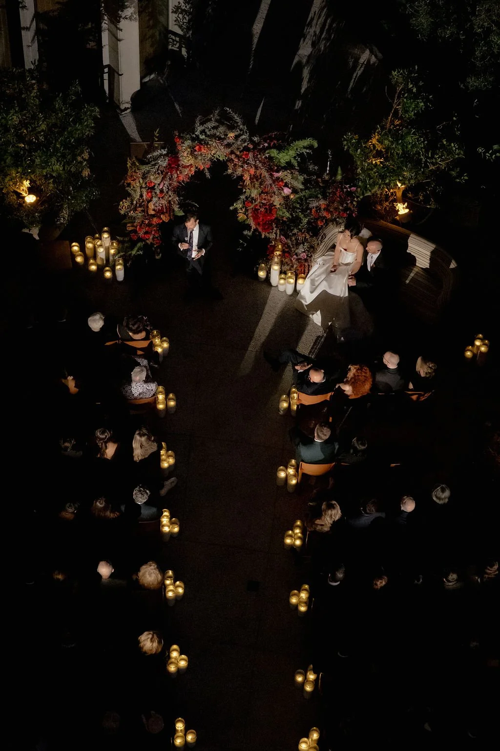 A wedding ceremony taking place outdoors at night, with the bride in a white gown and the groom seated next to her. An arch decorated with red and pink flowers and greenery frames the couple. Candles are placed along the aisle and surrounding the cer