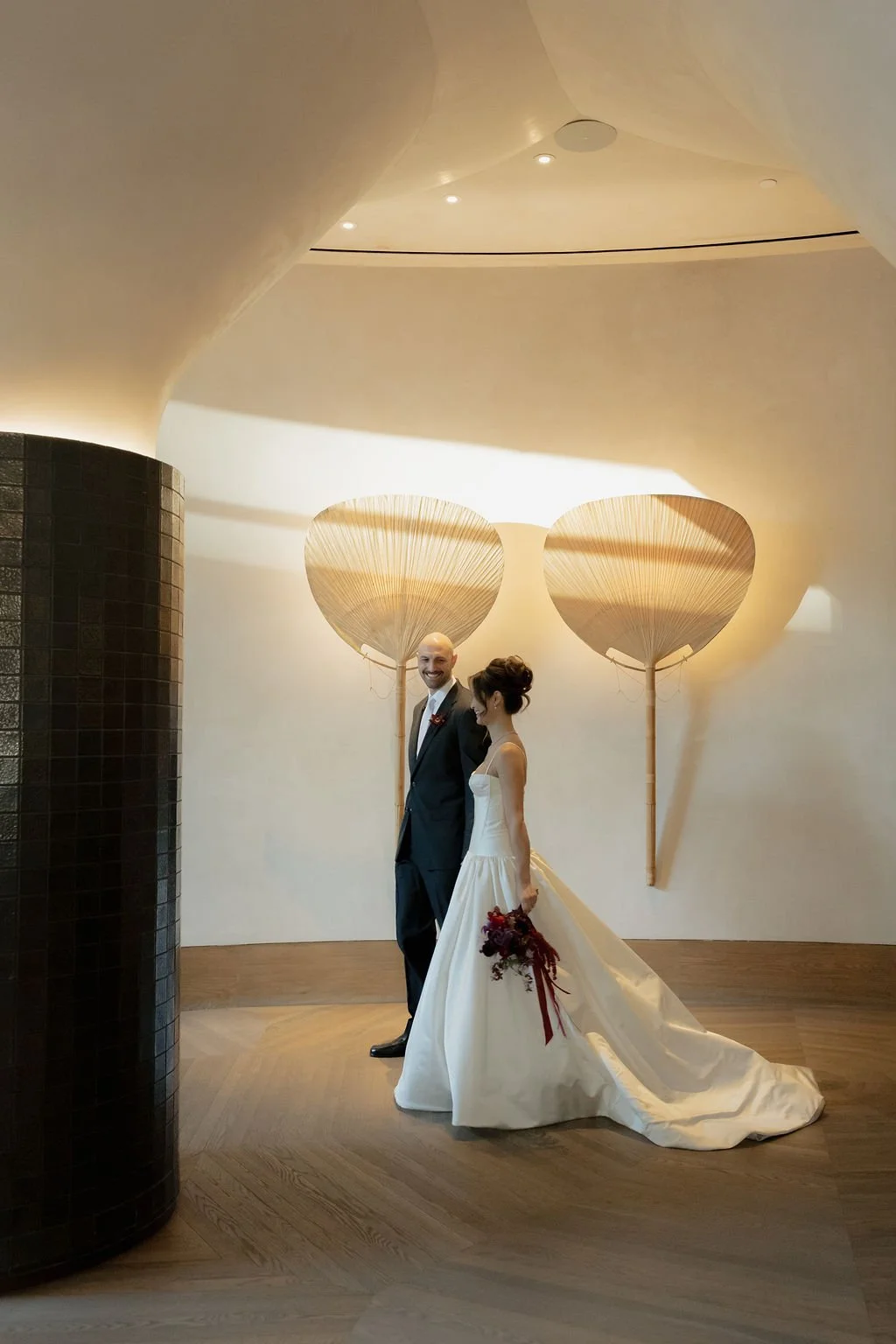 A bride and groom standing indoors, holding hands, with large decorative umbrellas on the wall behind them.