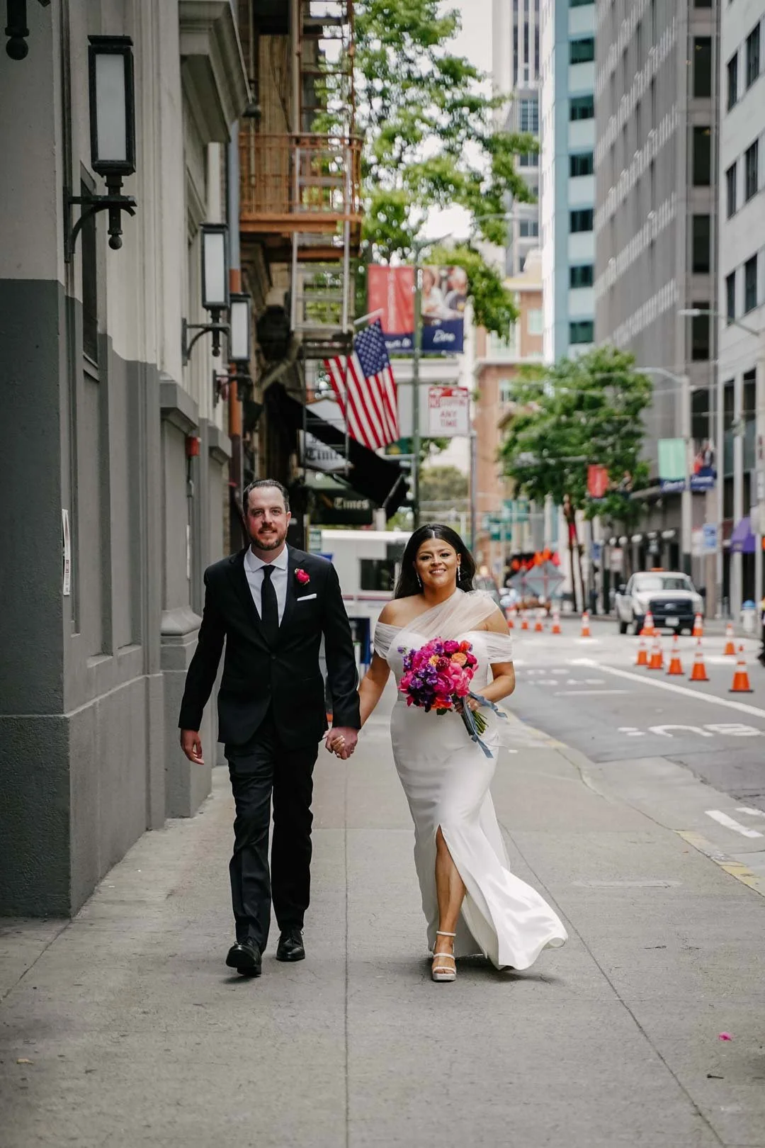A newlywed couple walking hand in hand down a city sidewalk, with the bride holding a bouquet of pink, purple, and red flowers, and wearing an off-the-shoulder white wedding gown, while the groom is dressed in a black suit and tie. City buildings and