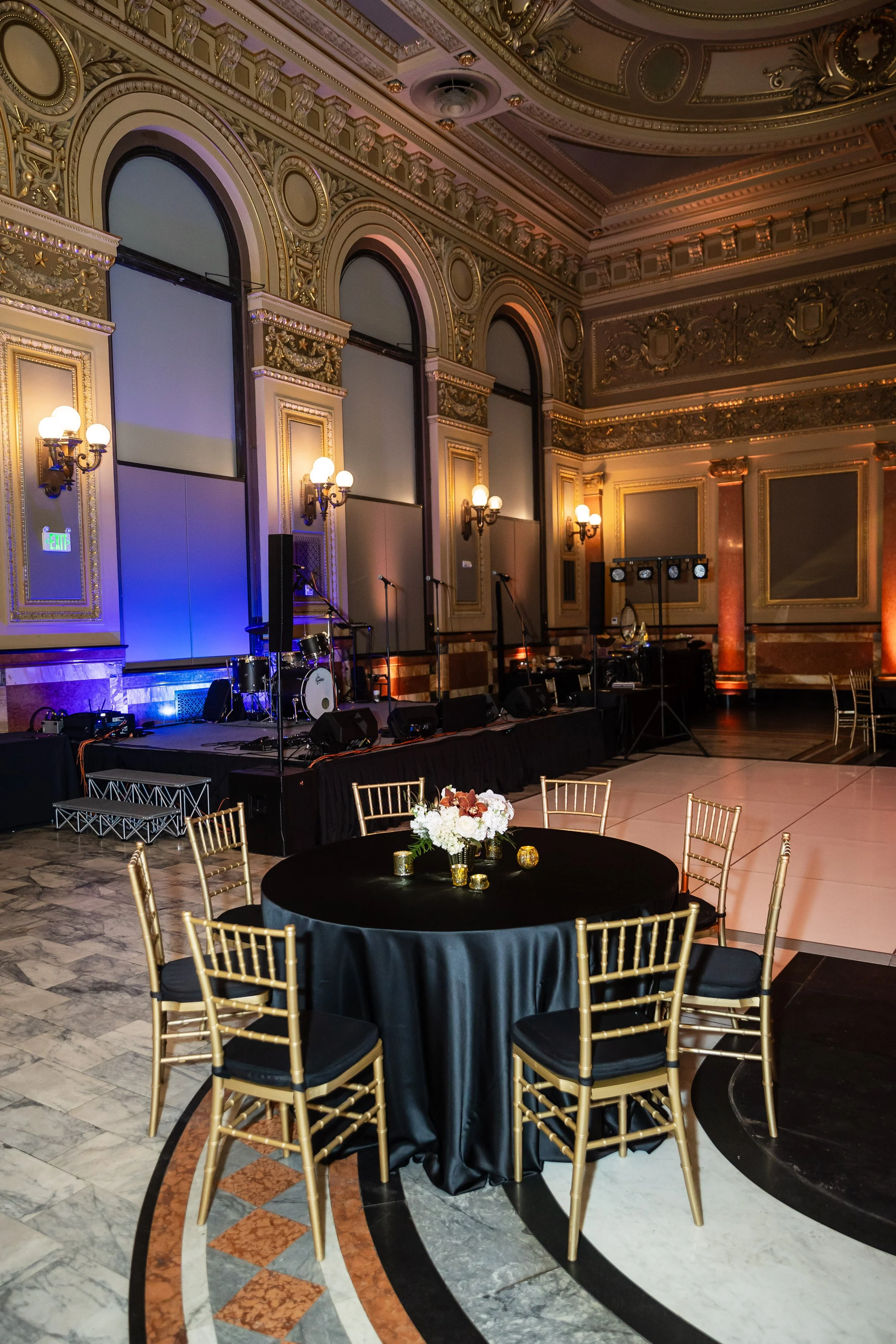 Elegant ballroom with a decorated round table with a floral centerpiece, a stage with musical instruments, and ornate gold and marble architectural details.