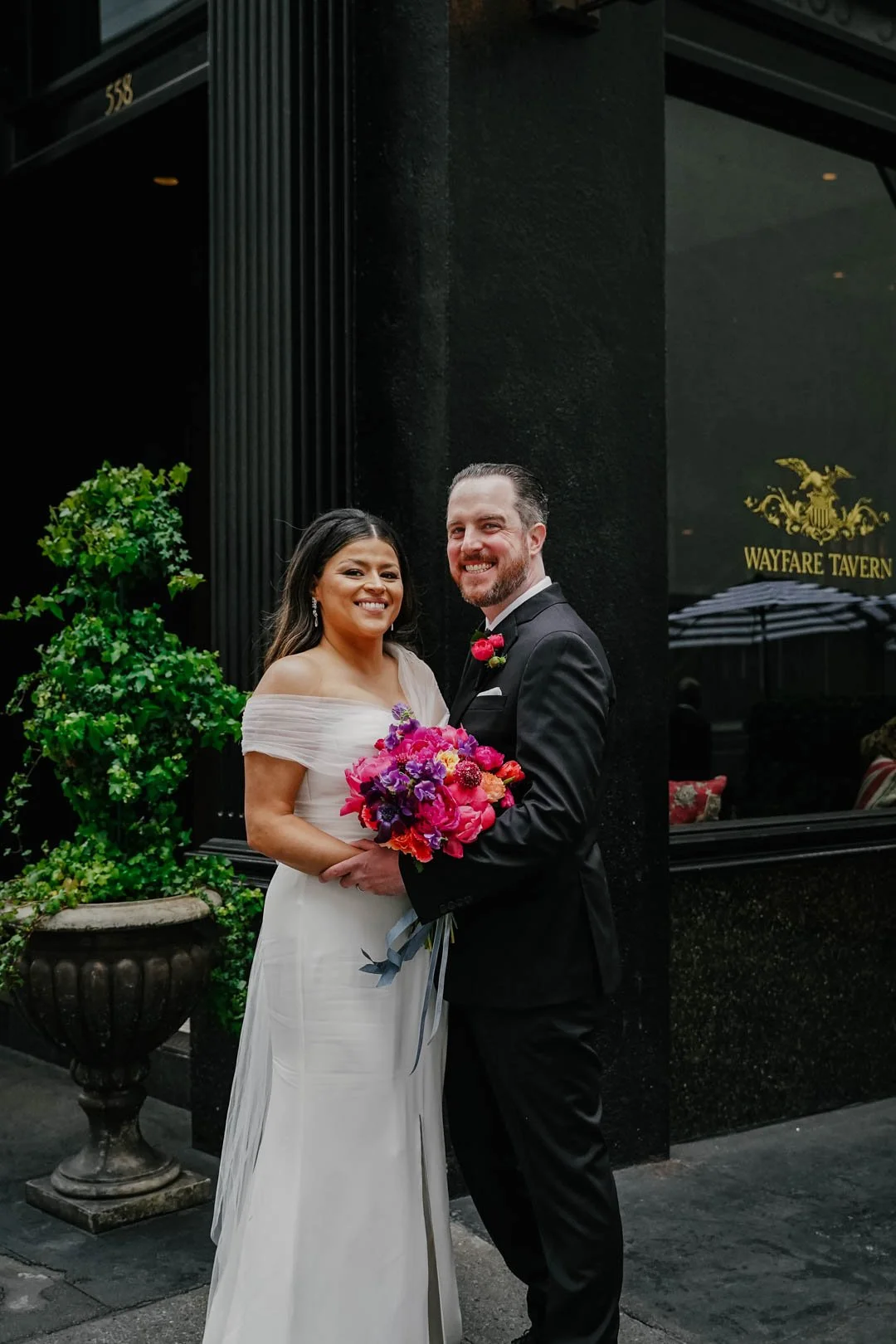 A newly married couple standing outside a restaurant called Warfare Tavern, smiling and holding a colorful bouquet of flowers.