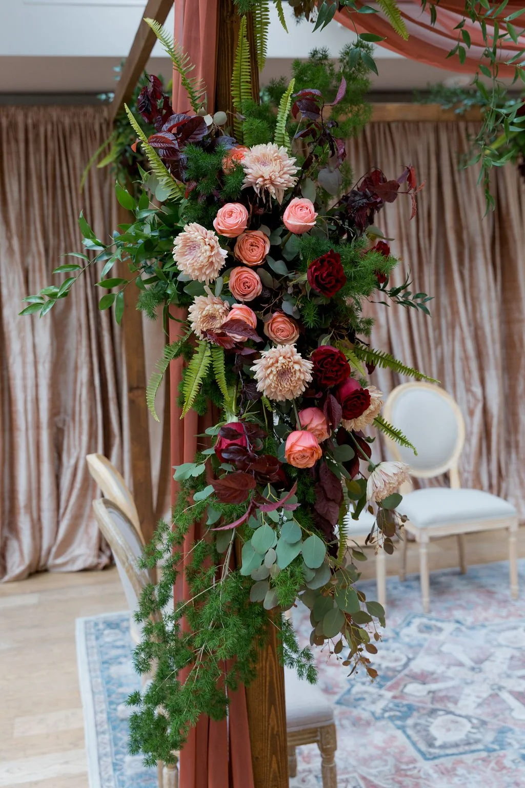 A floral arrangement featuring pink roses, cream dahlias, and red roses accented with green leaves and ferns, attached to a wooden structure, in an indoor setting with curtains and chairs in the background.