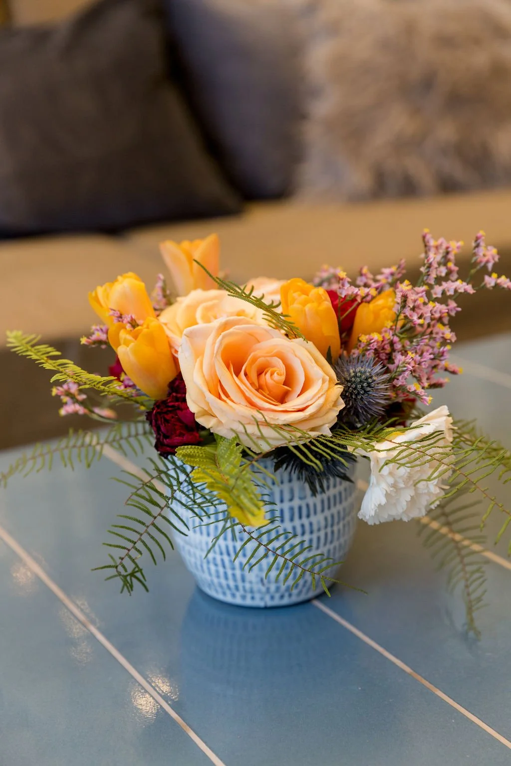 A bouquet of assorted flowers including pink roses, yellow tulips, purple thistle, and pink filler flowers in a white and blue patterned vase on a tiled table.