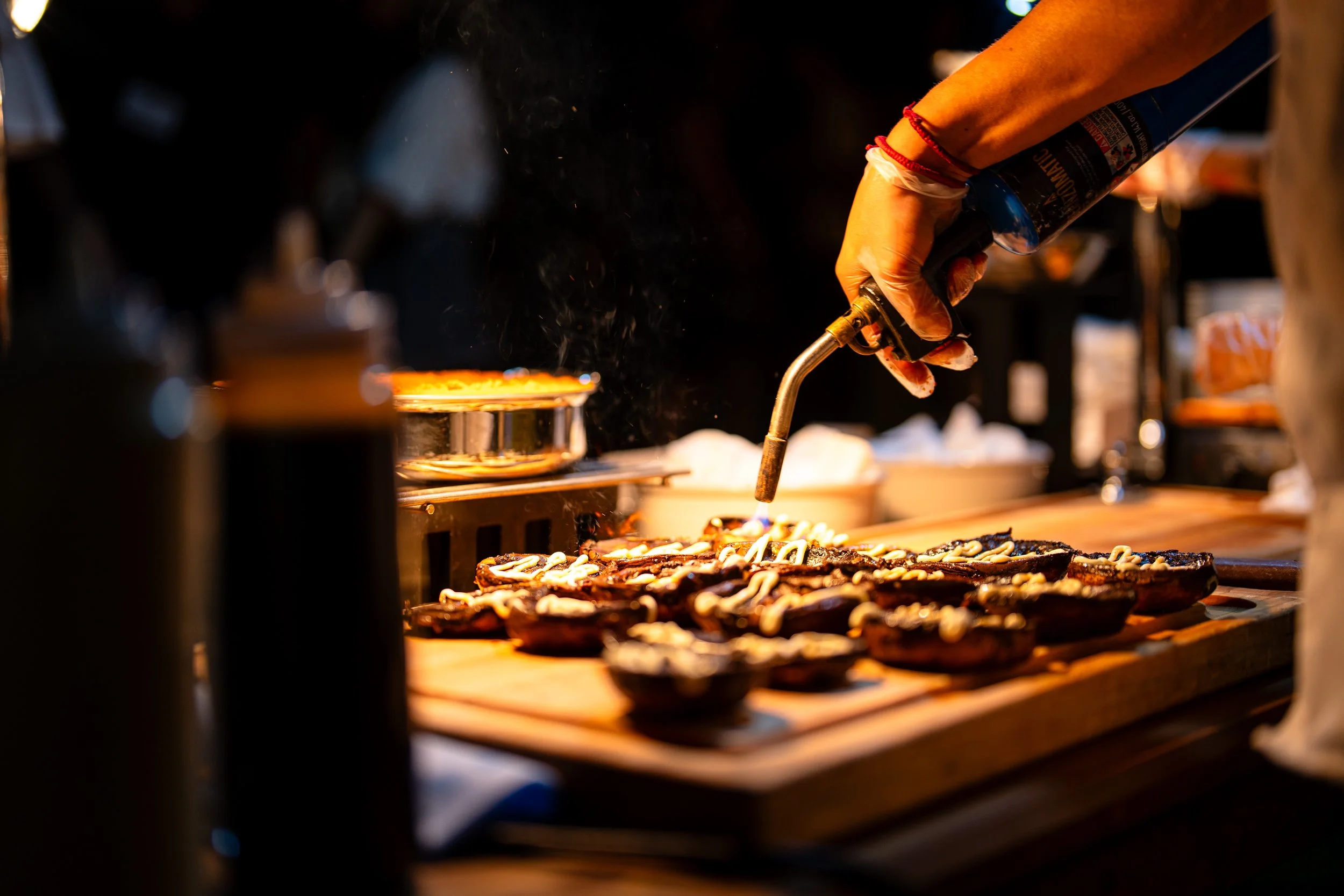 A person using a blowtorch to cook toppings on small pizzas or flatbreads on a wooden board at a food stand or kitchen.