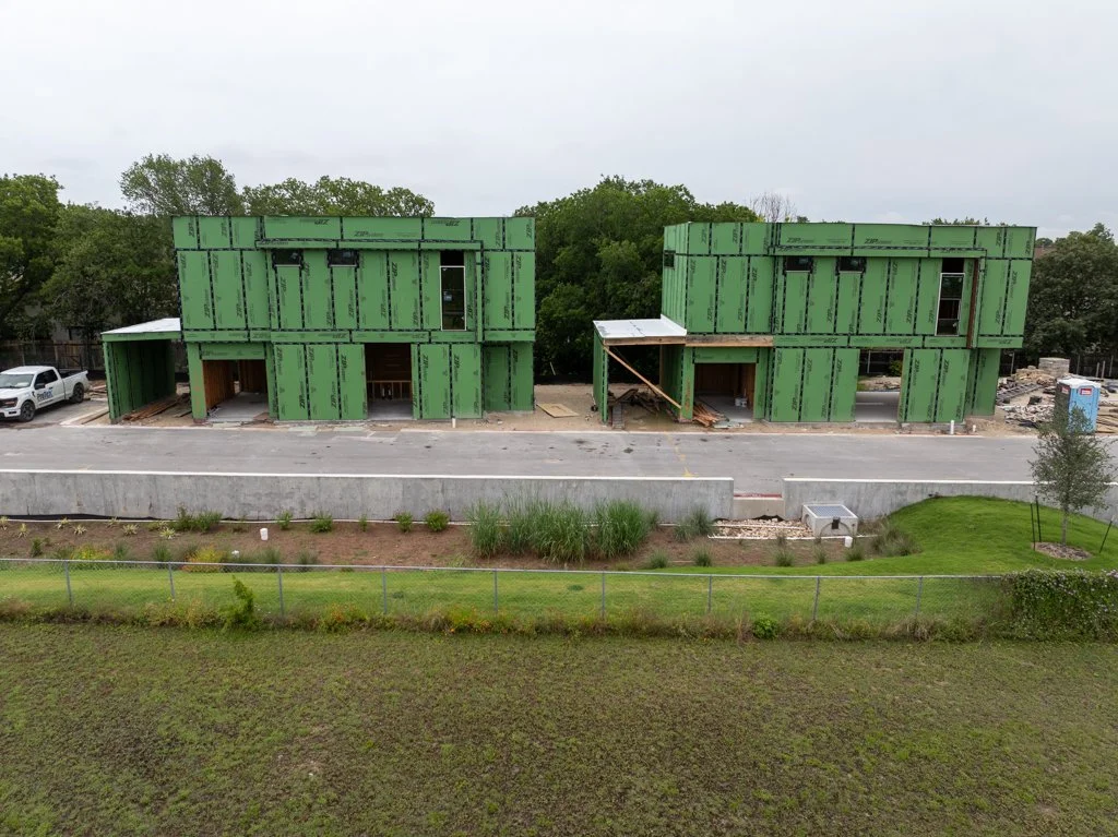 Two multi-story buildings under construction with green sheathing, situated behind a parking lot and green landscaped area.