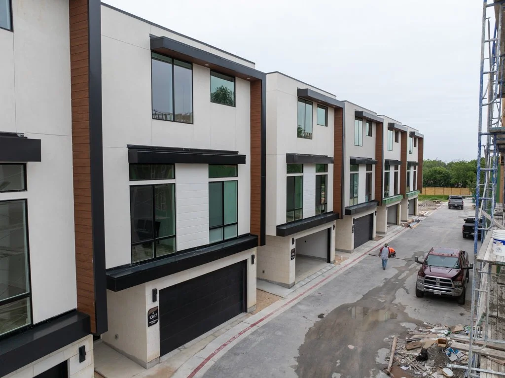 Newly constructed modern row of townhouses with large windows, black accents, and garage doors, under construction with scaffolding on the right, and a worker walking on the street.