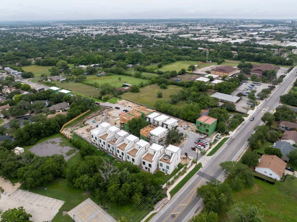 An aerial view of a residential area under construction, with a row of modern white townhouses, construction site, and surrounding greenery.